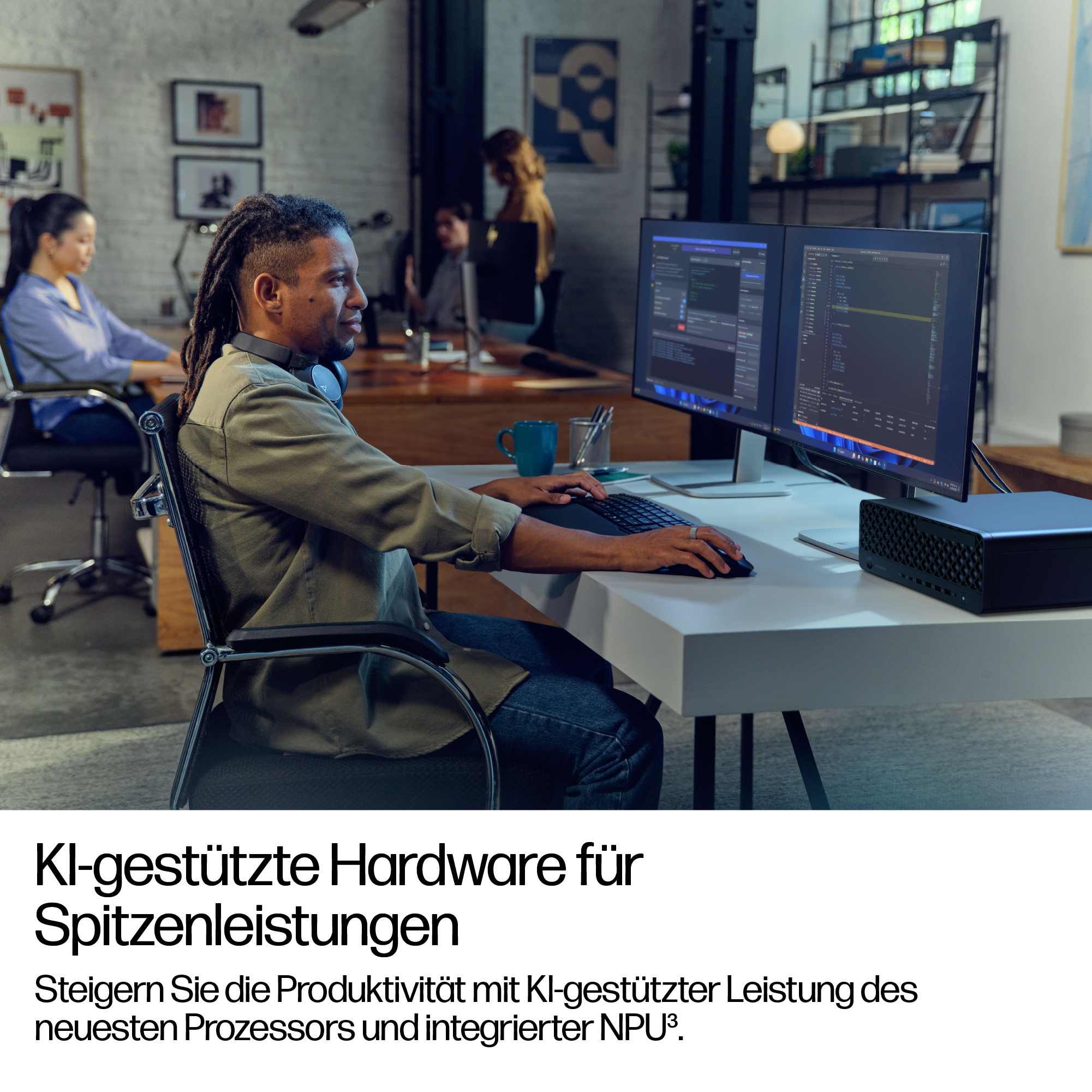 Ein Mann mit Dreadlocks arbeitet an einem Schreibtisch mit zwei Monitoren, einer Tastatur und einem schwarzen Computer. Andere Leute sind im Hintergrund.