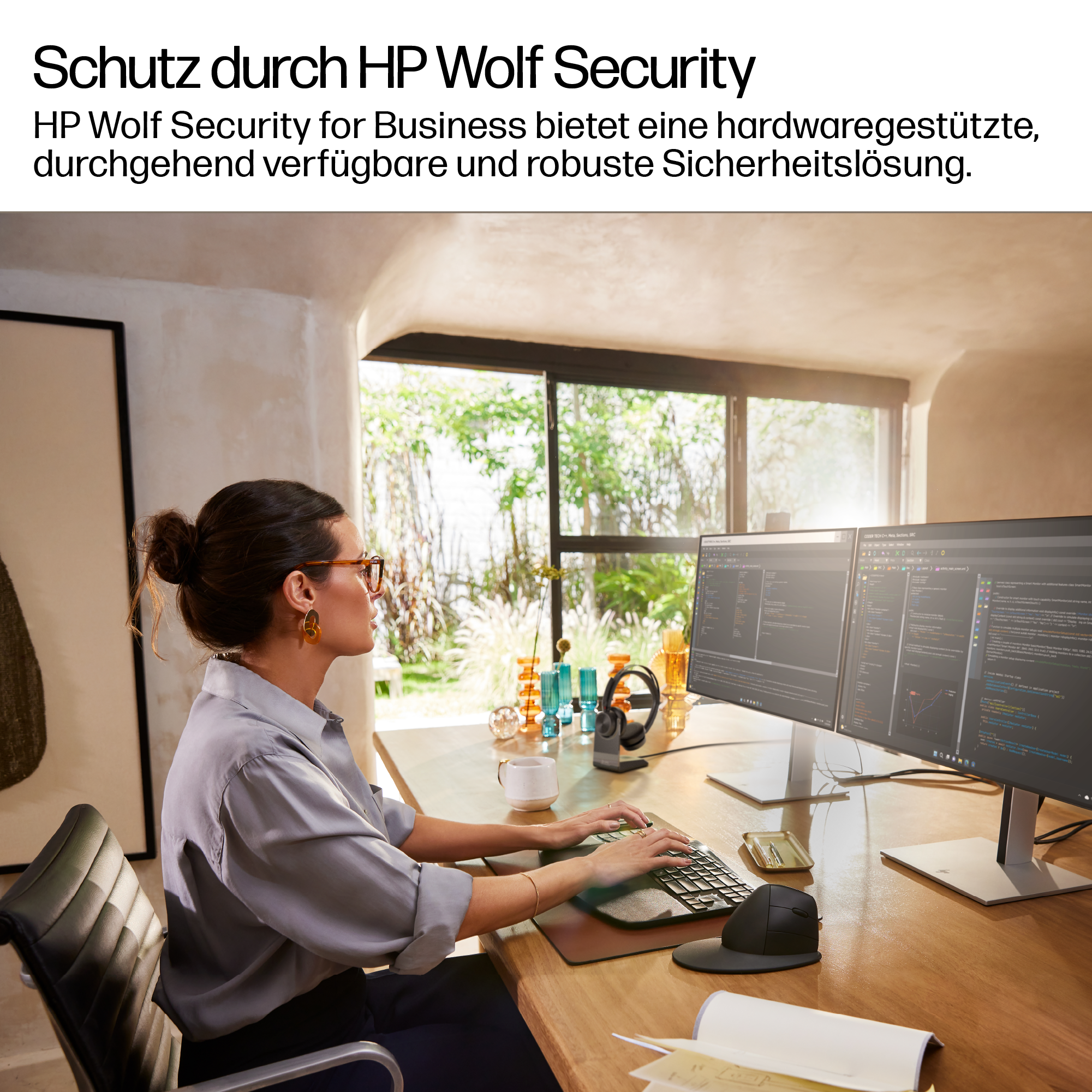 Eine Frau arbeitet an zwei Computerbildschirmen mit Code. Sie trägt eine Brille und sitzt an einem Holztisch. Im Hintergrund ist ein Fenster.