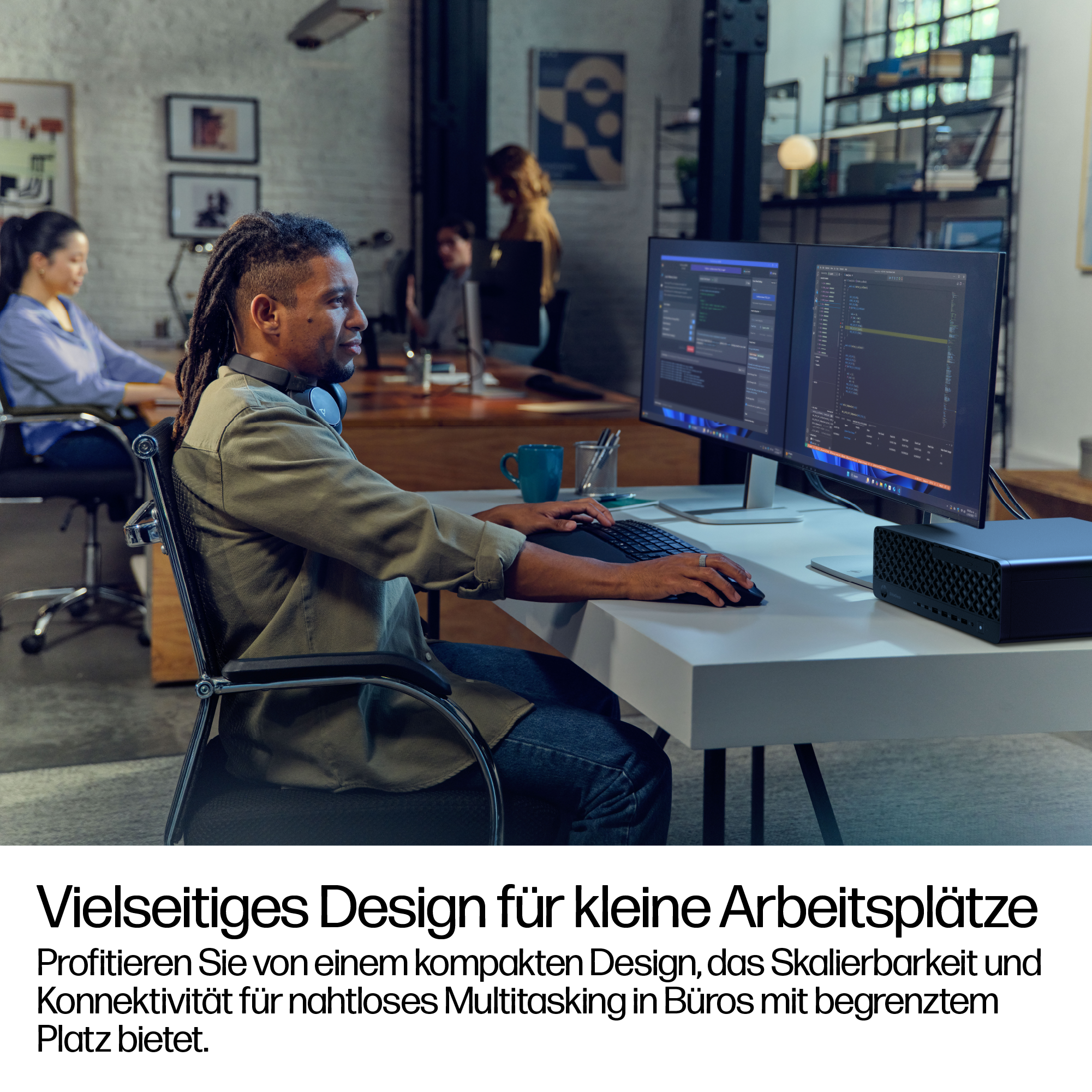 Ein Mann mit Dreadlocks arbeitet an einem Computer mit zwei Bildschirmen. Andere arbeiten im Hintergrund. Ein schwarzes Gerät steht auf dem Schreibtisch.