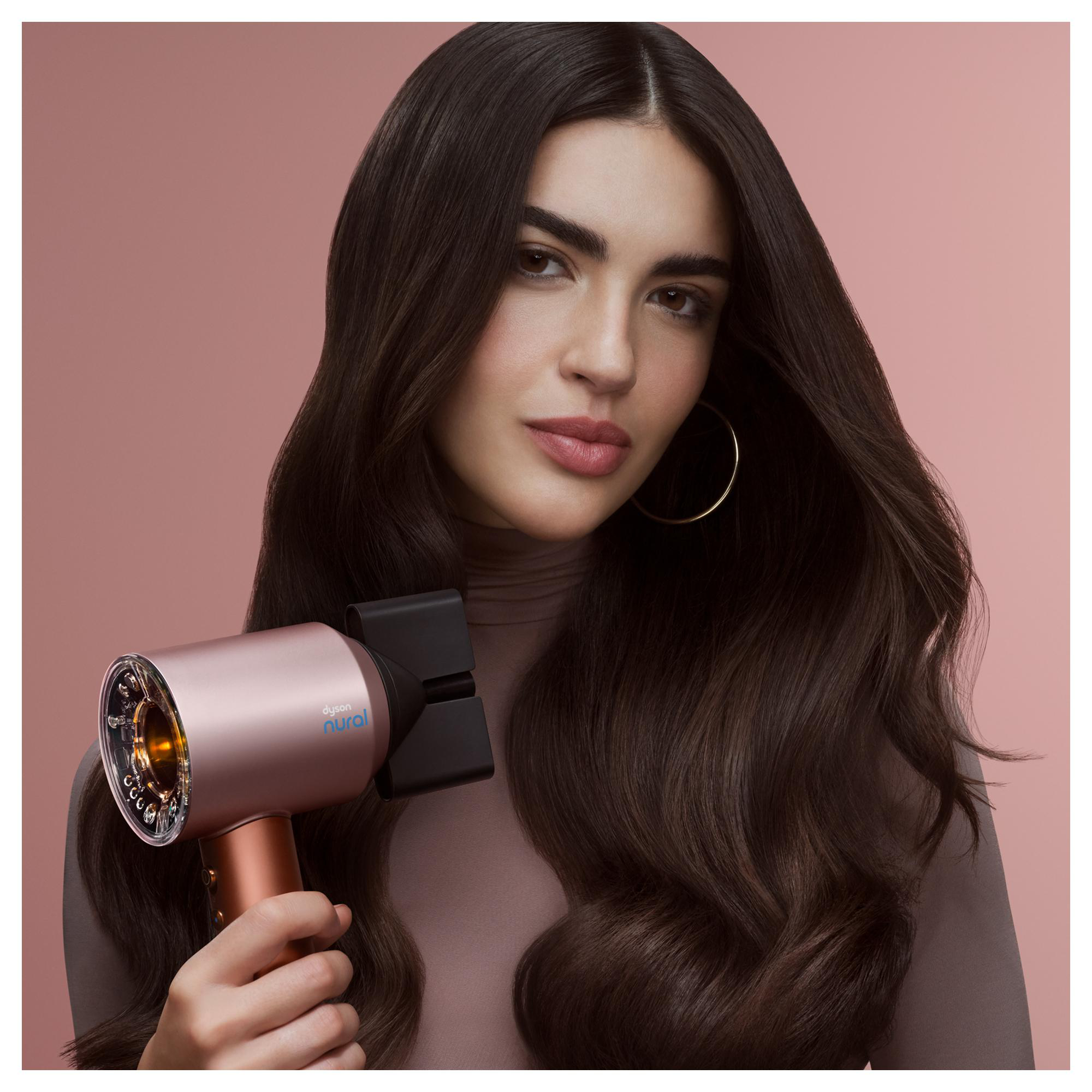 Eine Frau hält einen roségoldenen Haartrockner. Sie hat langes braunes Haar und trägt ein Oberteil in neutralem Ton. Der Hintergrund ist rosa.