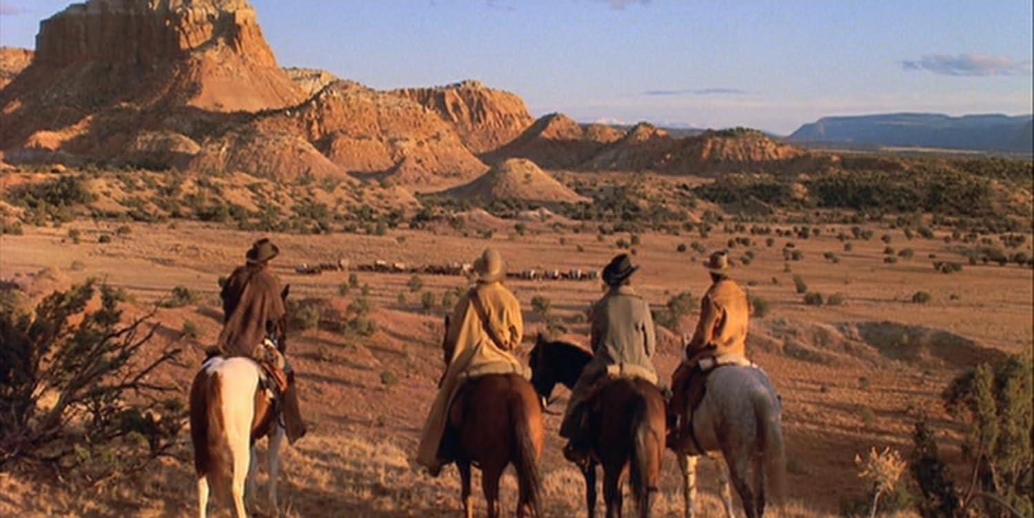Vier Cowboys auf Pferden reiten durch eine trockene Landschaft. Berge sind im Hintergrund. Vieh ist in der Ferne.
