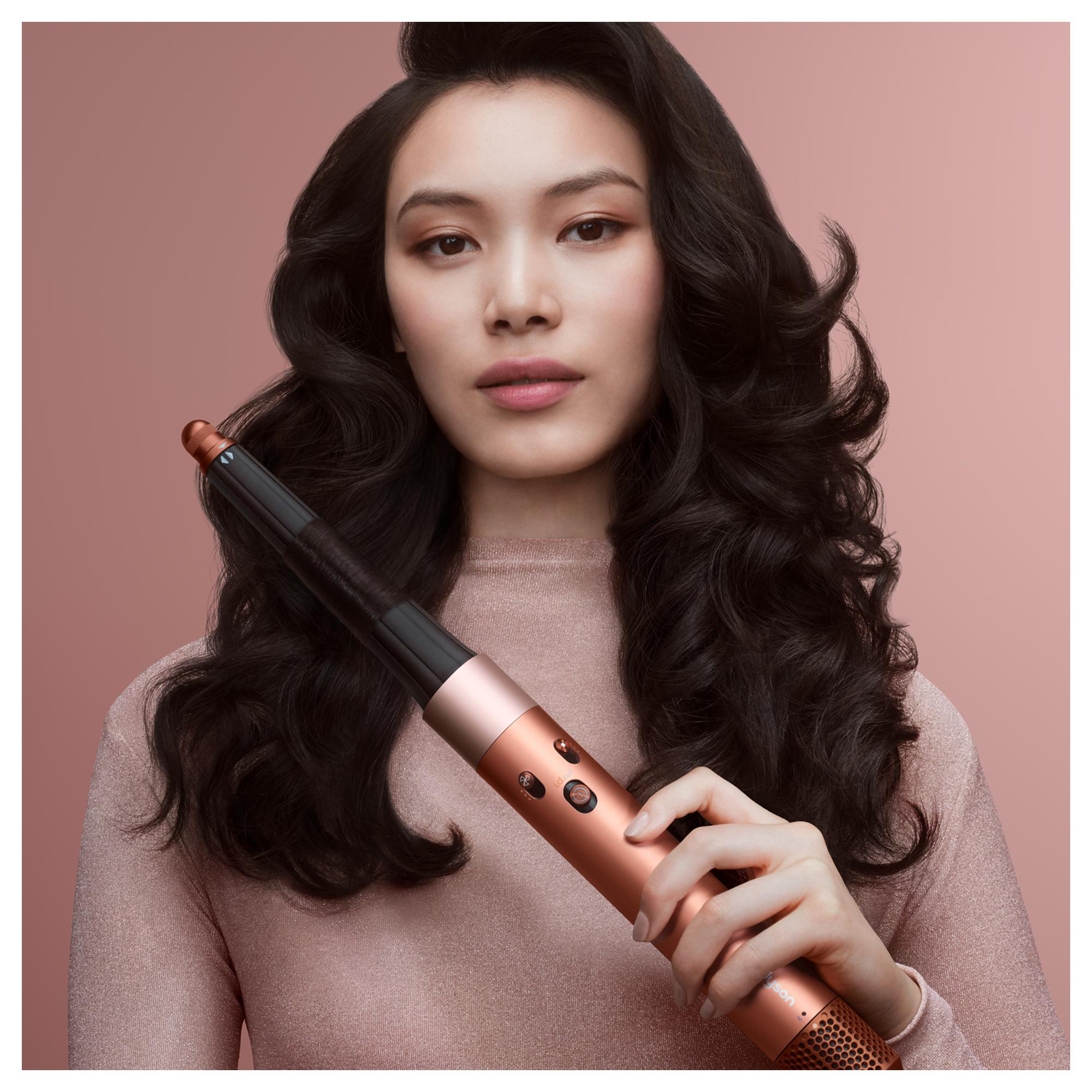 Frau hält einen kupferfarbenen Haarstyler, mit lockigem, dunklem Haar, vor rosa Hintergrund. Sie trägt ein rosafarbenes Oberteil. Der Styler ist nah an ihrem Gesicht.