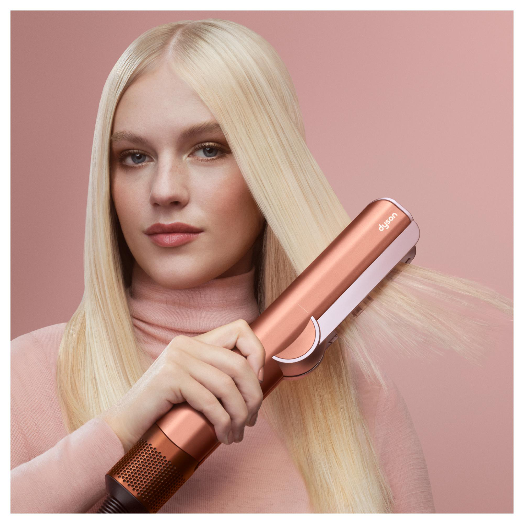 Frau mit blondem Haar benutzt ein Dyson-Haarglätteisen. Sie trägt einen rosa Rollkragenpullover, der Hintergrund ist rosa.