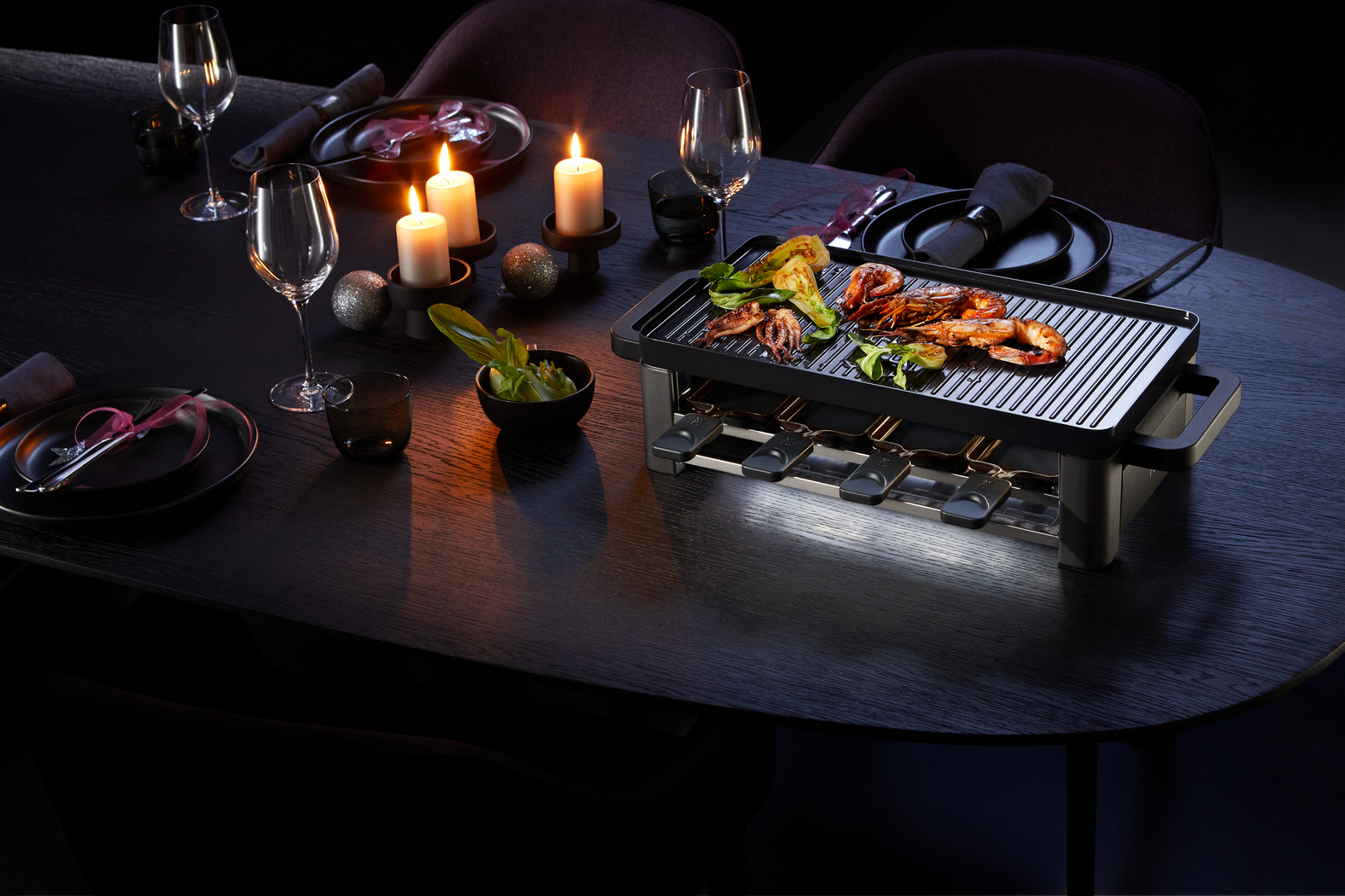 Tisch gedeckt mit Raclette-Grill, Essen, Kerzen und Weingläsern. Dunkle, stimmungsvolle Atmosphäre.