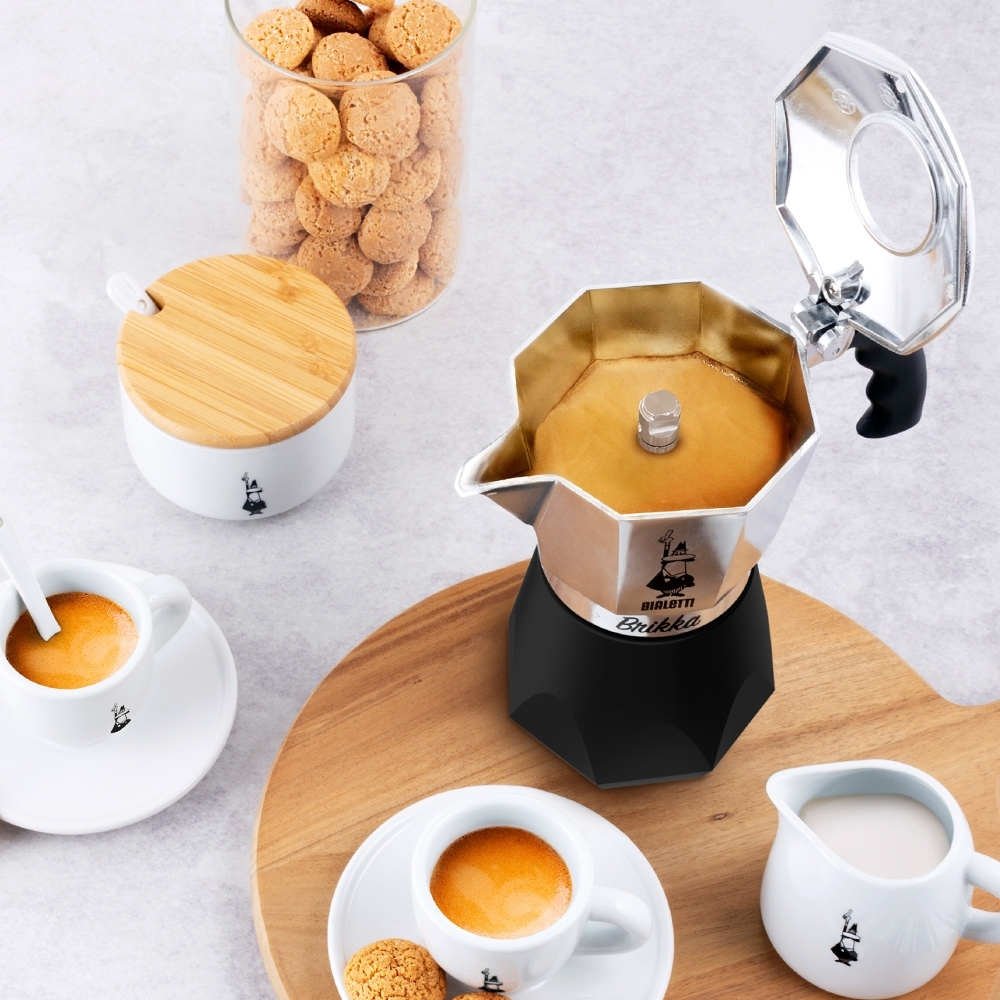 Ahşap bir tahta üzerinde Moka potu, espresso fincanları, kurabiyeler, şekerlik ve sütlük bulunan bir kahve düzeni.