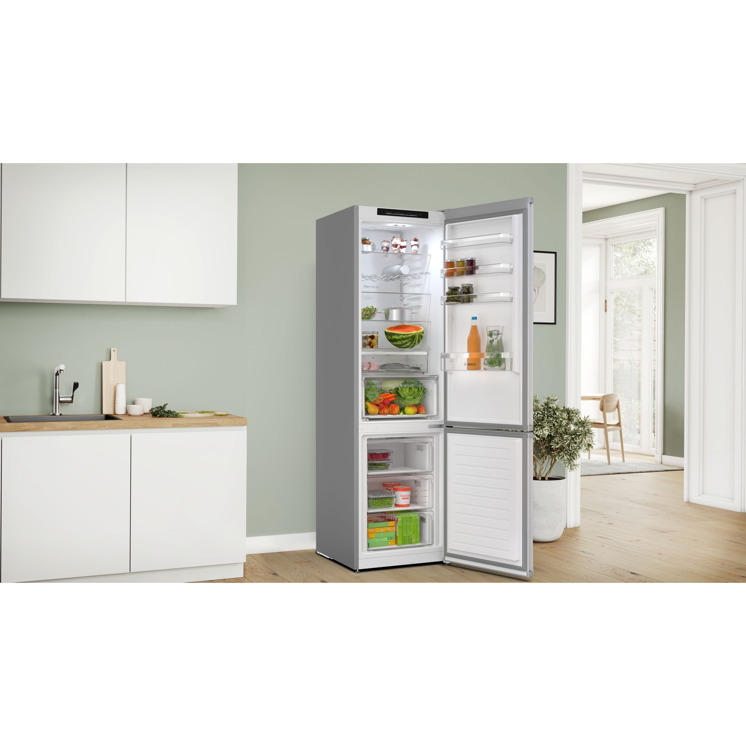 Un frigorifero argentato con la porta aperta piena di cibo. Una cucina con lavandino, piano di lavoro e altri articoli da cucina è sullo sfondo.