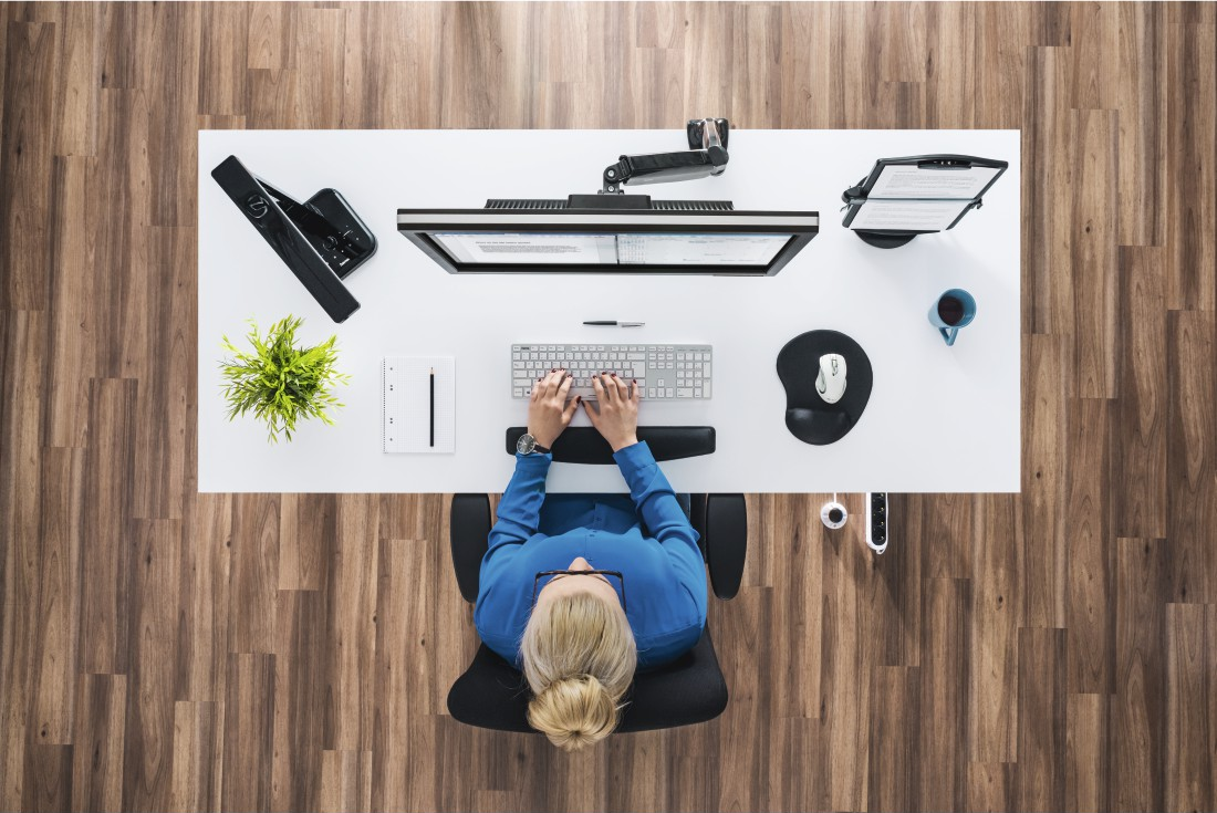 Vue aérienne d'une femme travaillant à un bureau blanc. Elle tape sur un clavier. Un moniteur, une souris, une tasse et une plante sont visibles.