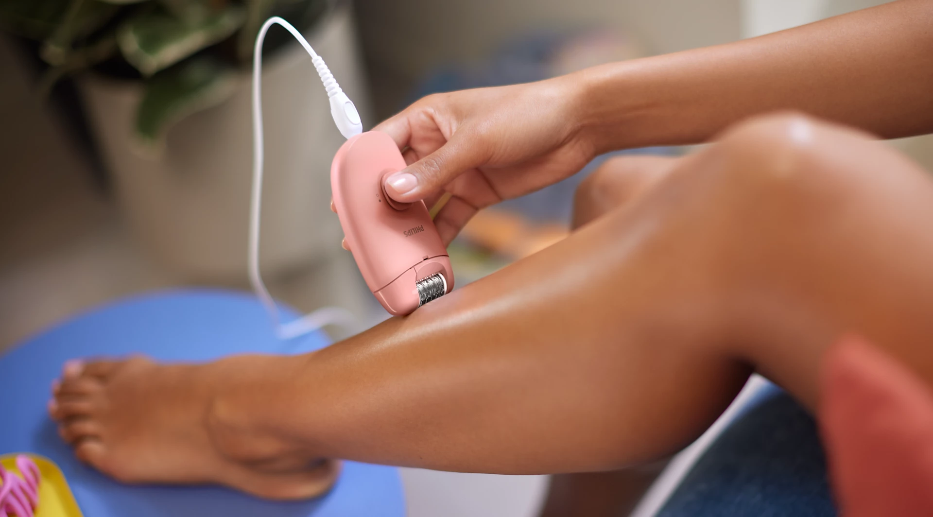 Une personne utilise une épilateur rose sur sa jambe. La jambe est au centre, avec le pied et l'épilateur visibles. Une plante est en arrière-plan.