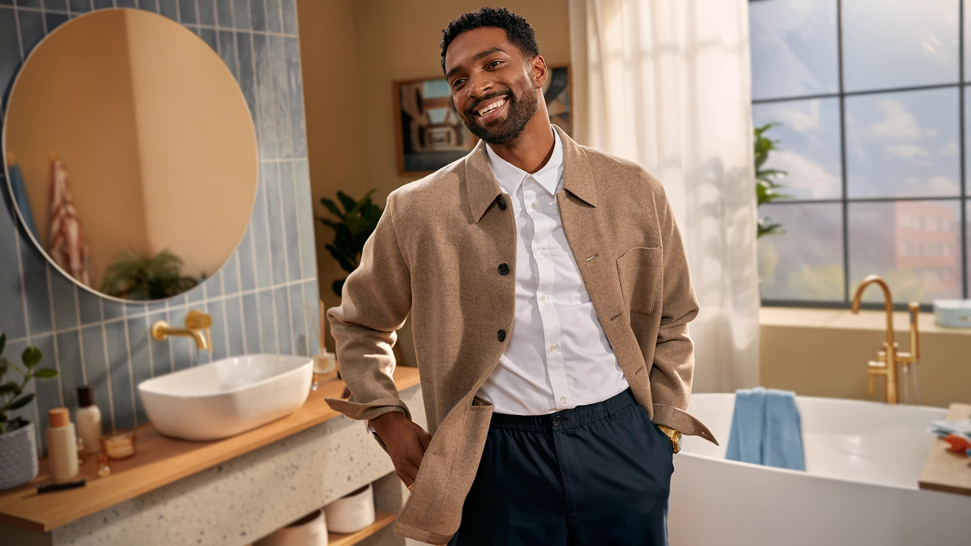 Un uomo sorride in un bagno. Indossa una camicia bianca e una giacca marrone. Sullo sfondo ci sono un lavandino, uno specchio e una vasca da bagno.
