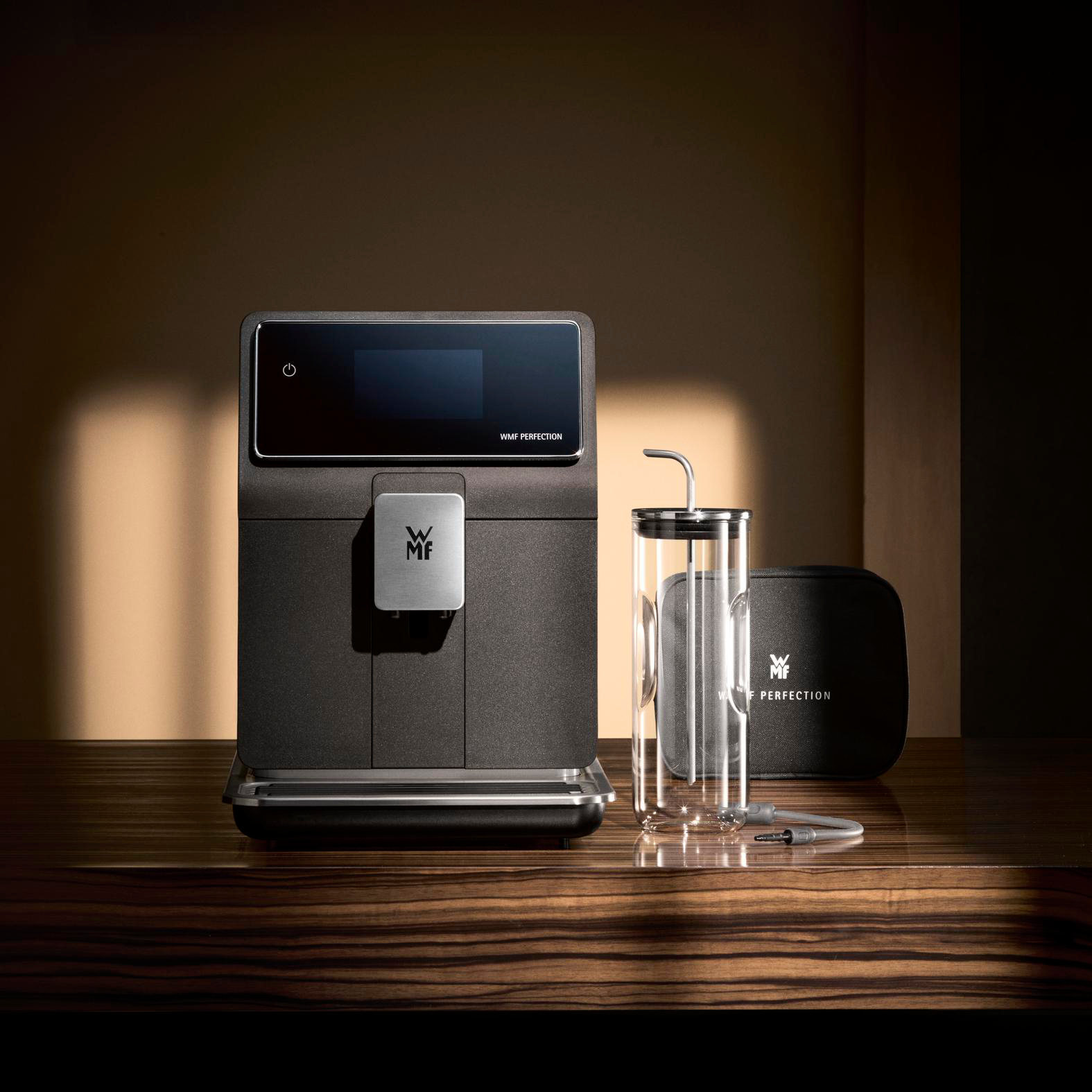 Eine schwarze WMF Perfection Kaffeemaschine, ein Glasbehälter, eine schwarze Tasche und ein silbernes Kabel auf einer Holzoberfläche.