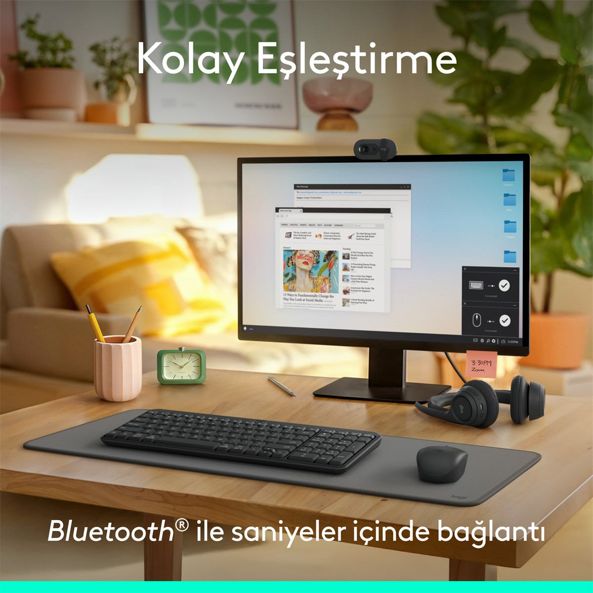 Bir monitör, klavye ve fare bulunan bir masa. Monitörün üzerinde bir web kamerası var. Bir saat ve kulaklık mevcut. Arka plan bitkileri içerir.