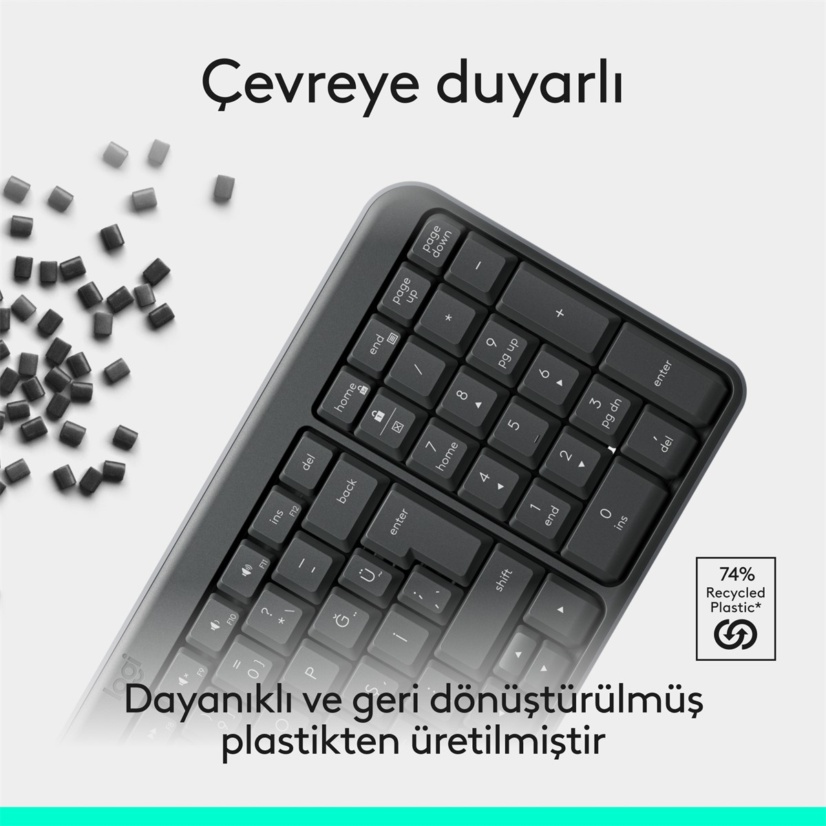 Beyaz tuşları ve sayıları olan koyu gri bir klavye. Metin '74% Geri Dönüşümlü Plastik' yazıyor. Arka planda dağılmış siyah plastik peletler.