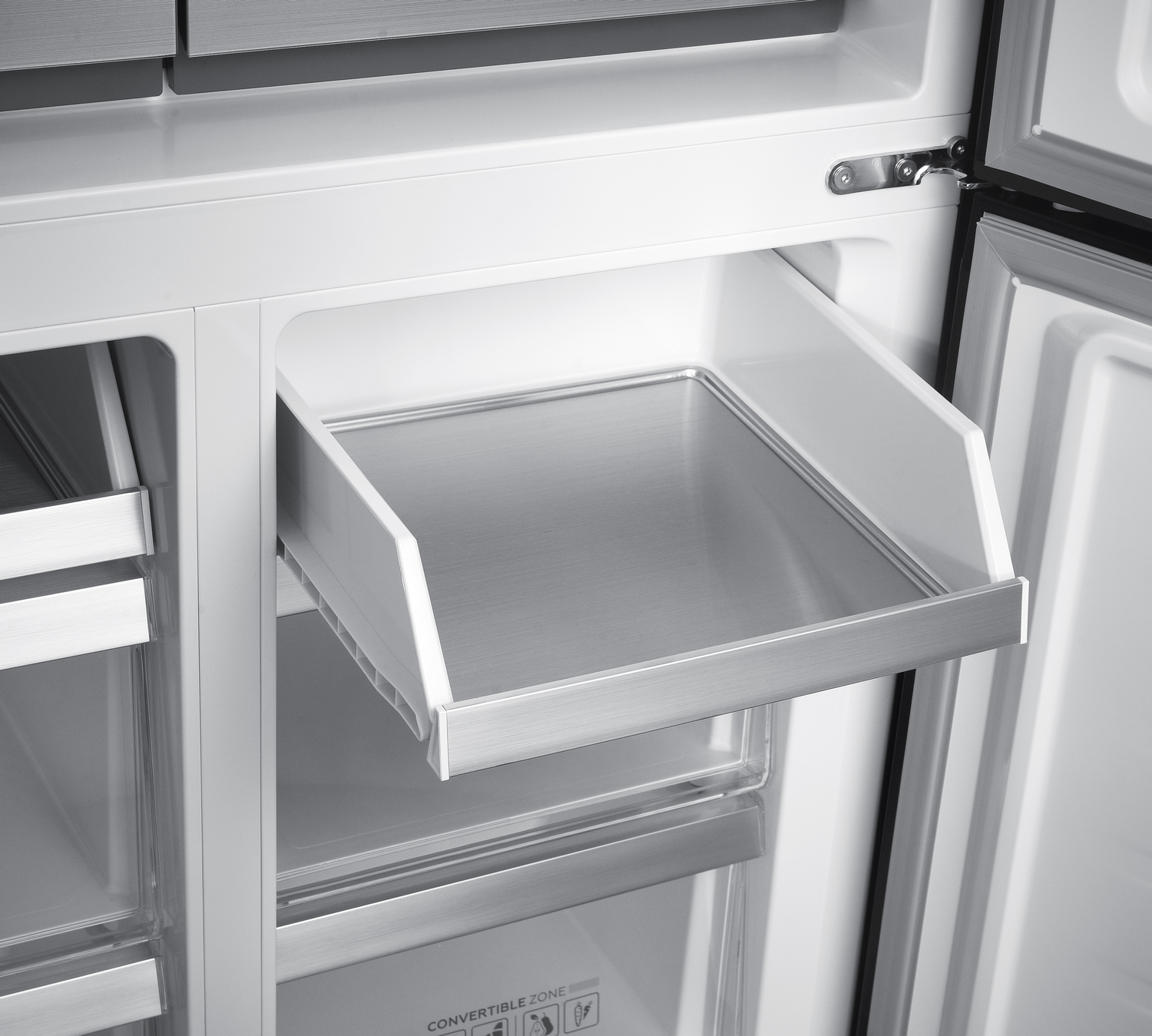 Dentro de un refrigerador, un cajón abierto con superficie de acero inoxidable. Interior blanco con puerta parcialmente abierta. Etiqueta de zona convertible visible.