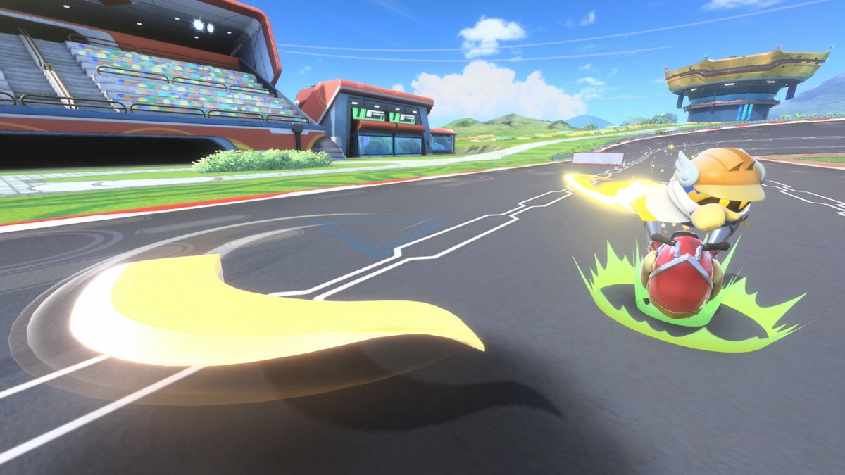 A character on a red vehicle with a yellow trail on a racetrack. Buildings and a stadium are in the background under a blue sky.