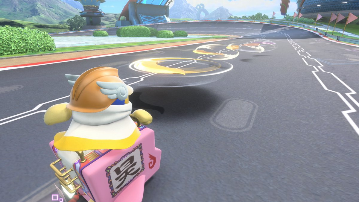 A character in a kart, wearing a helmet, races on a track. Mountains and buildings are in the background. The kart has a pink body.
