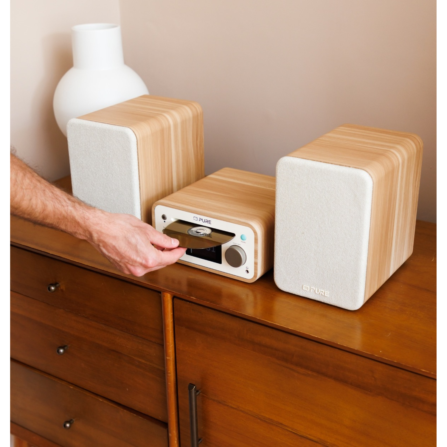 A person inserts a CD into a wooden stereo. Two speakers and the stereo sit on a wooden cabinet. A white vase is in the background.