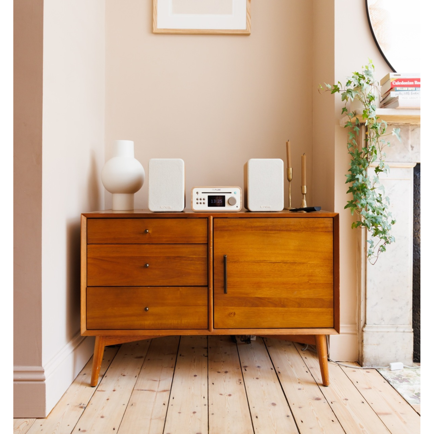 A wooden cabinet with a white vase, speakers, and a radio. A plant grows on the right. The room has wooden floors.