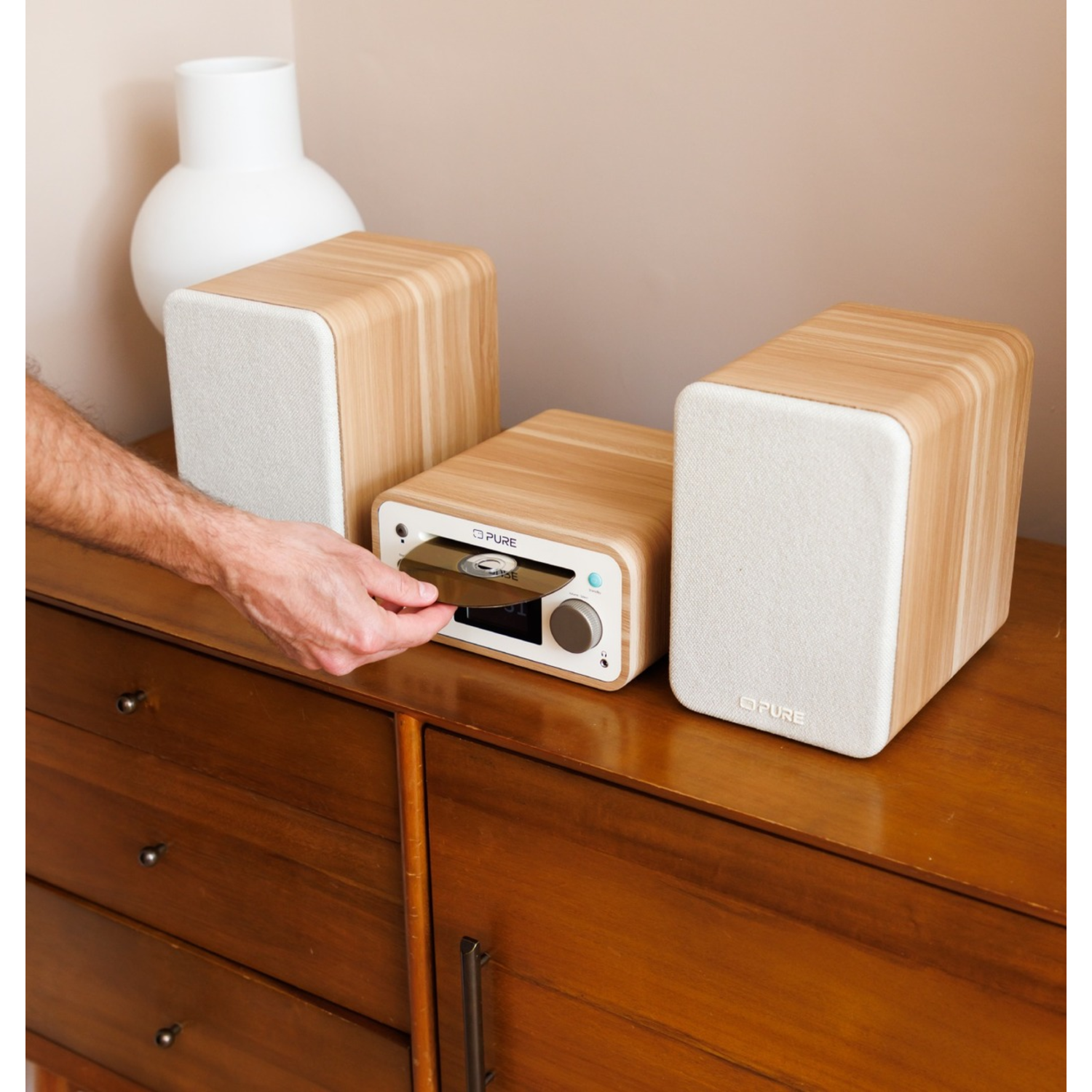 A person inserting a CD into a wooden stereo system. The stereo sits on a wooden cabinet, with a white vase behind it.