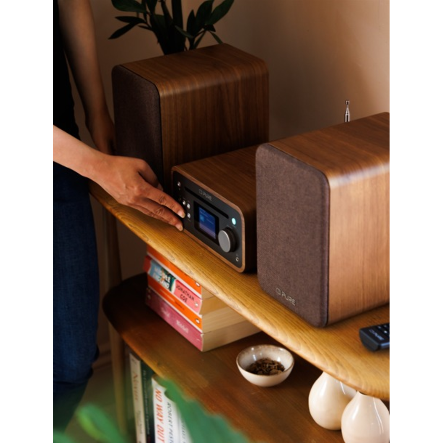 A person's hand adjusts a wooden stereo system. Books, small bowl, and vases are on a wooden shelf.