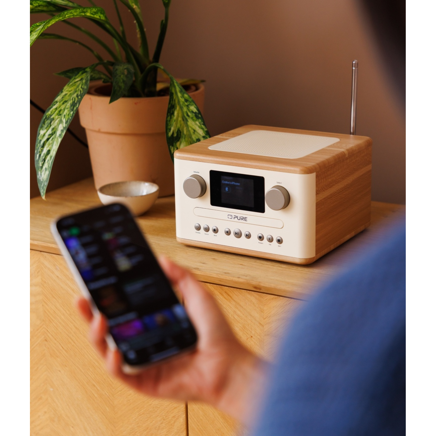 A person holds a phone near a radio on a wooden surface. A plant in a pot is on the left. The radio has a display.
