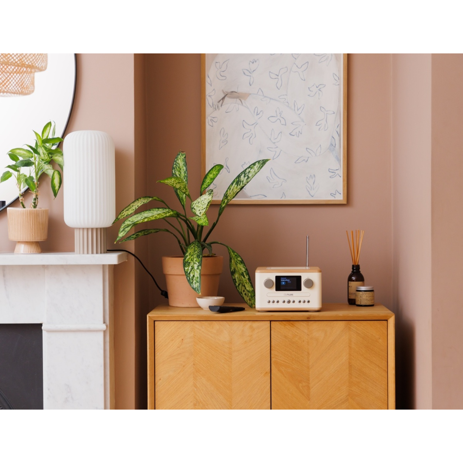 A room with a wooden cabinet, plants, a radio, and a painting on the wall. The wall is painted in a soft pink color.