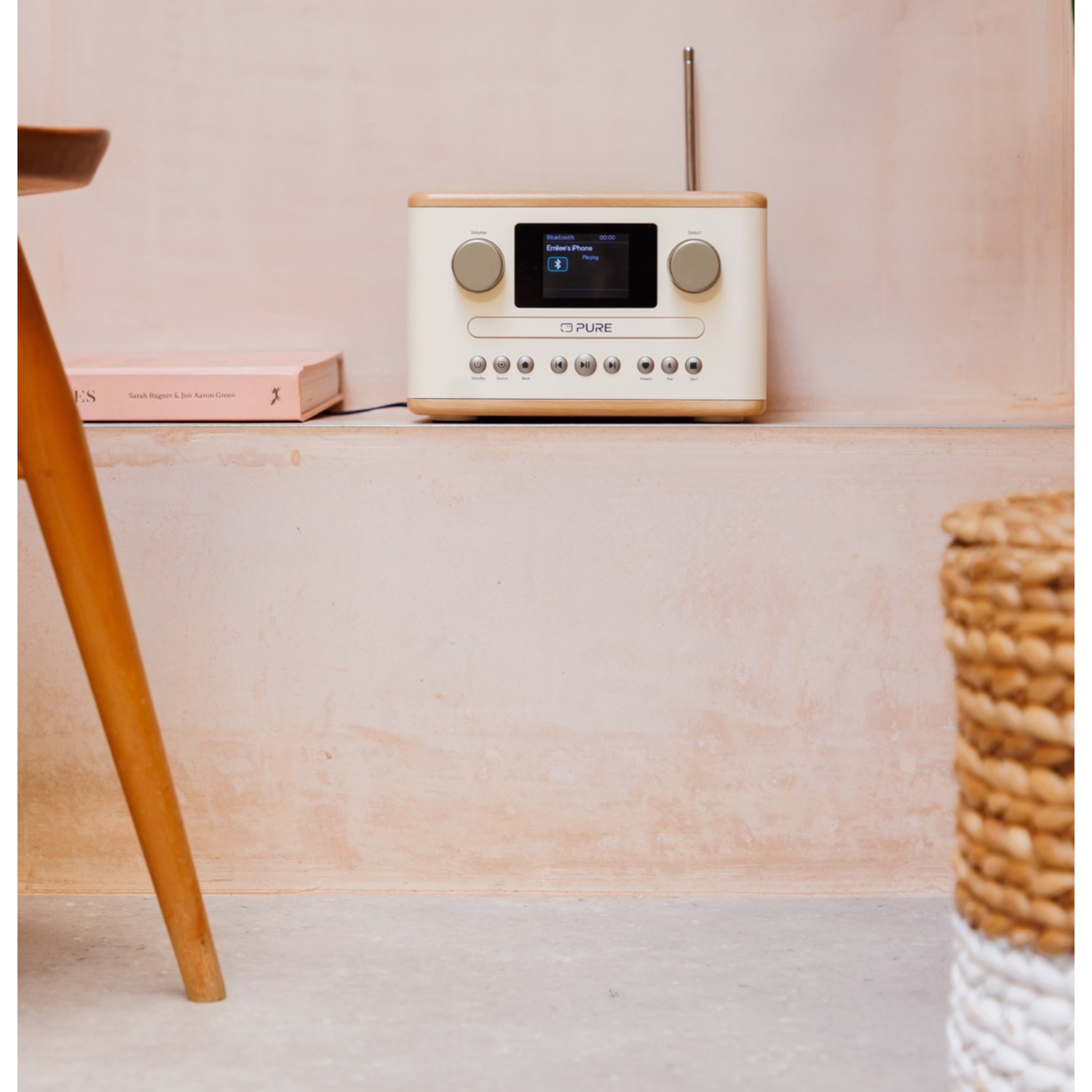 A cream radio with a screen is on a ledge with a book and a wooden stool leg. A woven basket is in the background.