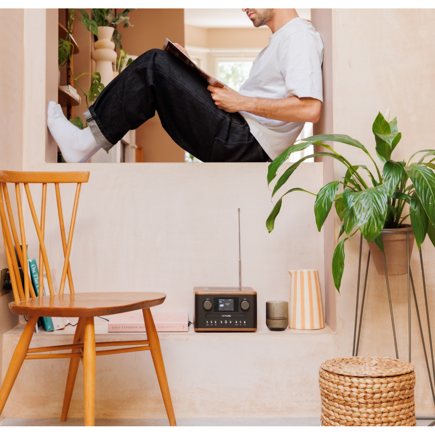 Man reading on ledge. Wooden chair, radio, plant, and wicker basket. Neutral colors.