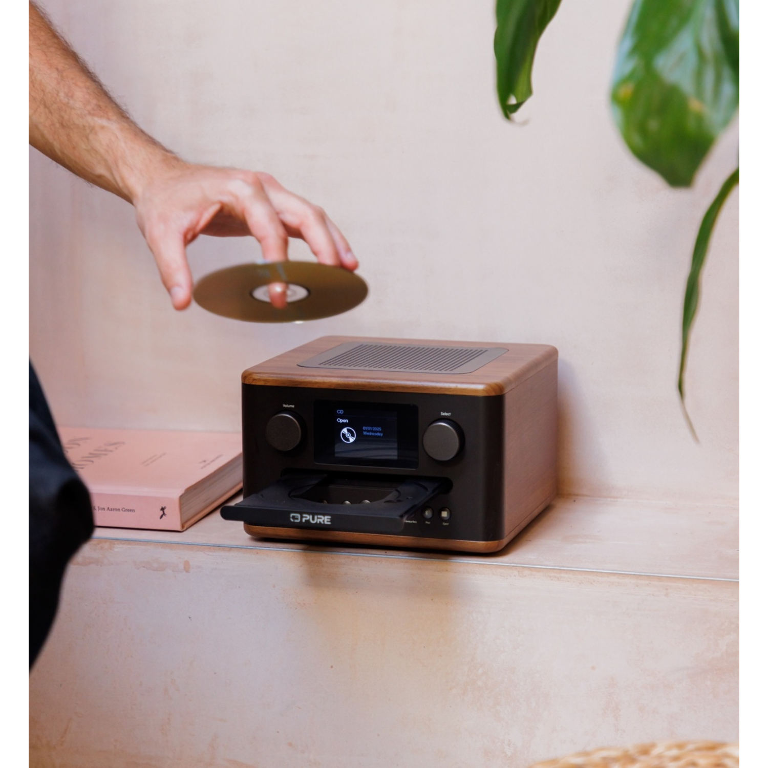 A hand inserting a CD into a wooden radio. A book and a plant are on the sides. The radio has a display.