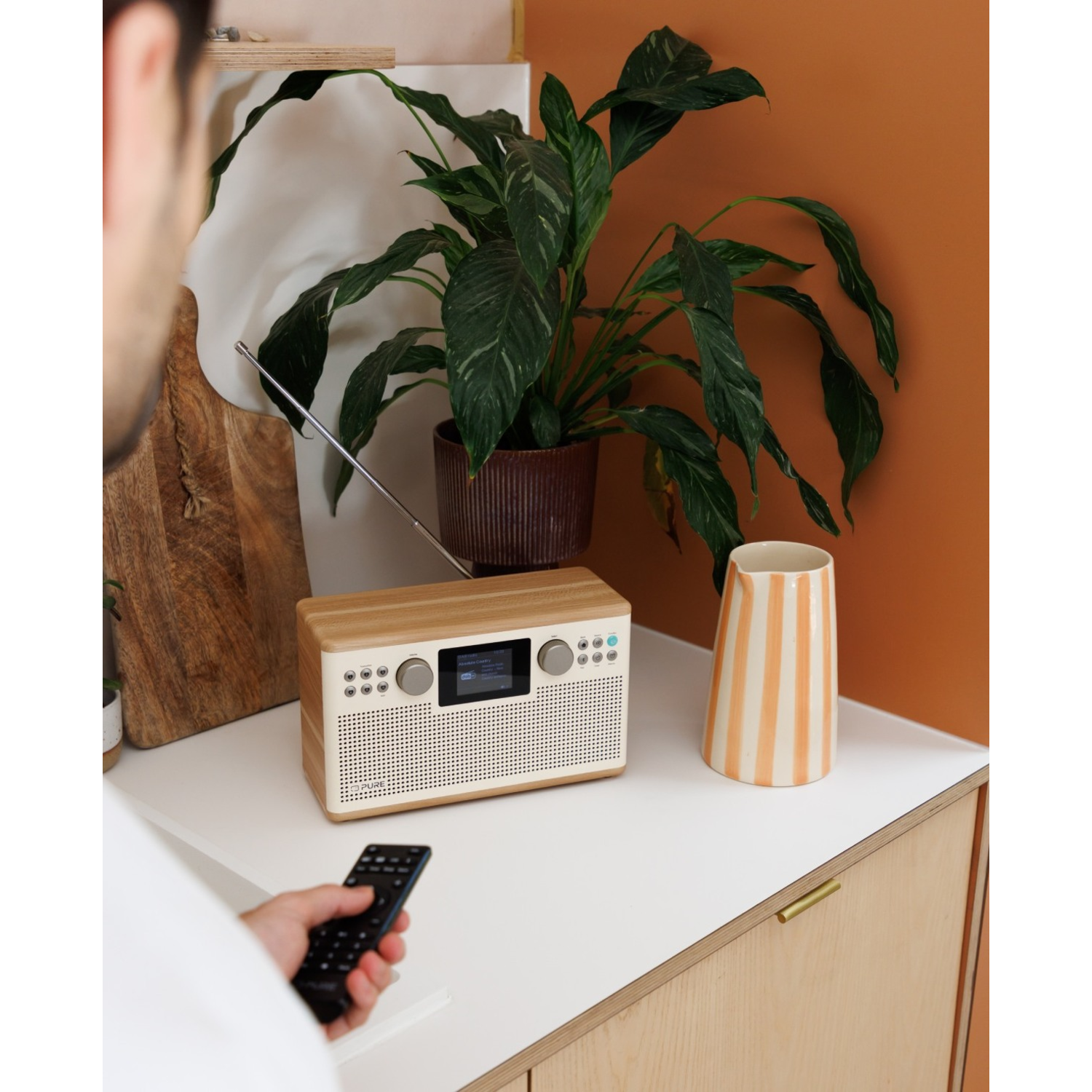 Eine Person hält eine Fernbedienung auf ein Radio. Eine Pflanze, eine Vase und ein Holzbrett stehen auf einer weißen Oberfläche.