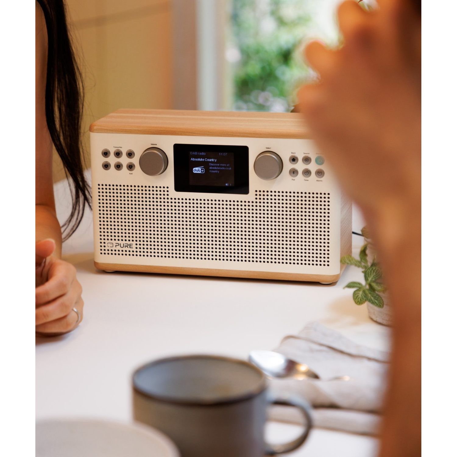 Ein Radio mit der Aufschrift 'Absolute Country' auf dem Bildschirm steht auf einem Tisch mit Tassen, Löffeln und Personen. Der Hintergrund ist unscharf.