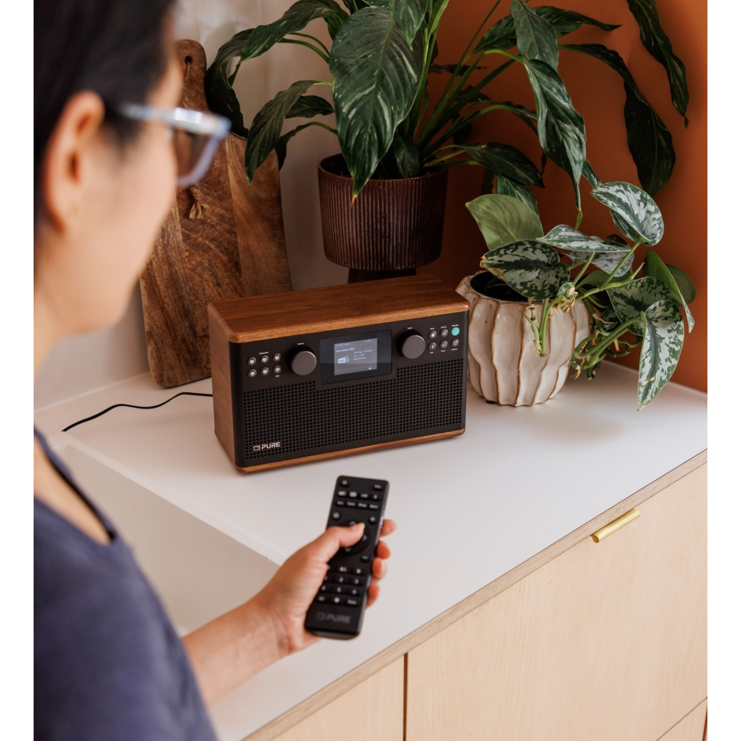 Eine Person mit Fernbedienung bedient ein Radio. Pflanzen in Töpfen sind im Hintergrund. Das Radio steht auf einer weißen Oberfläche.