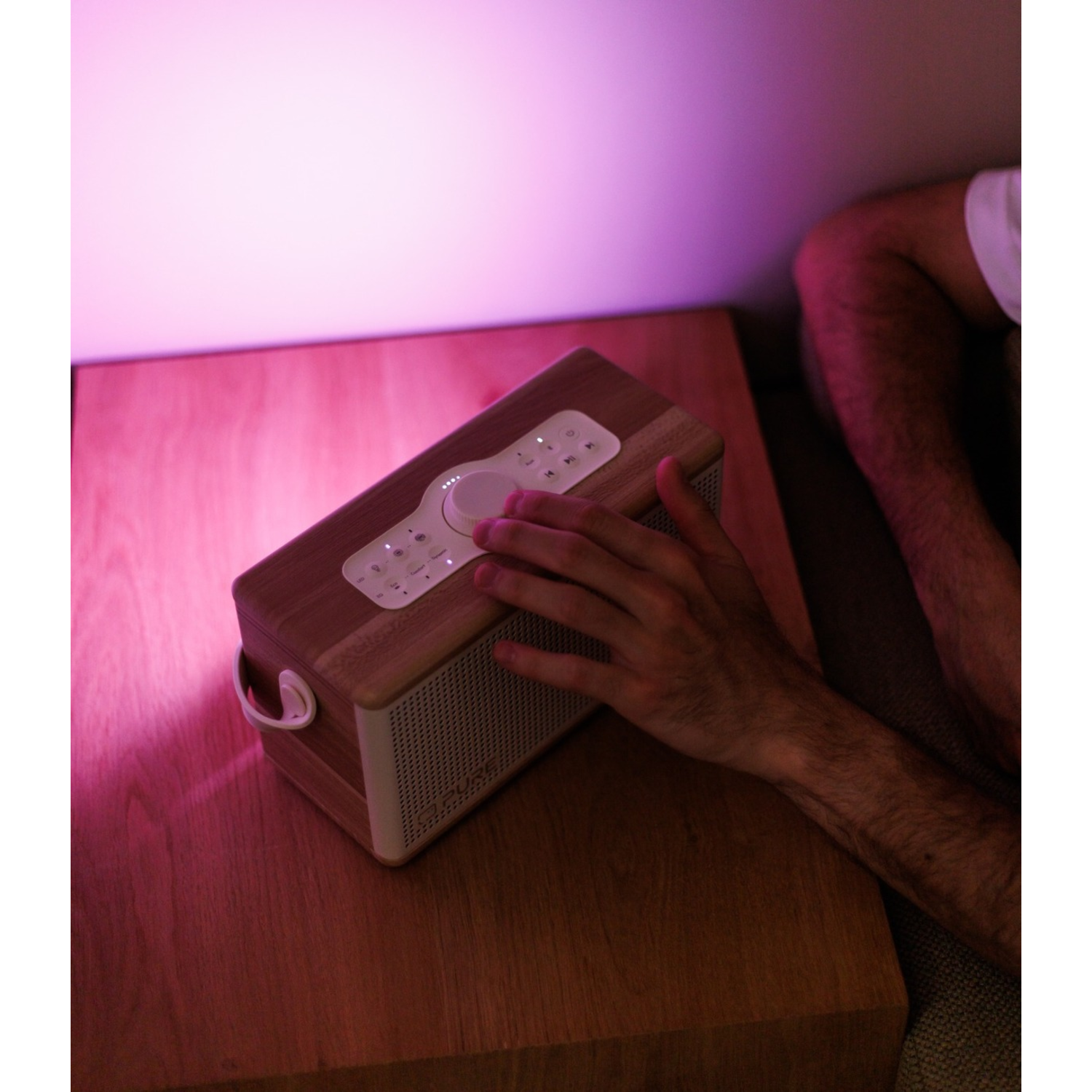 Die Hand einer Person stellt ein Holzradio auf einem Nachttisch ein. Rosa Licht beleuchtet den Raum, mit Teilansicht der Person.