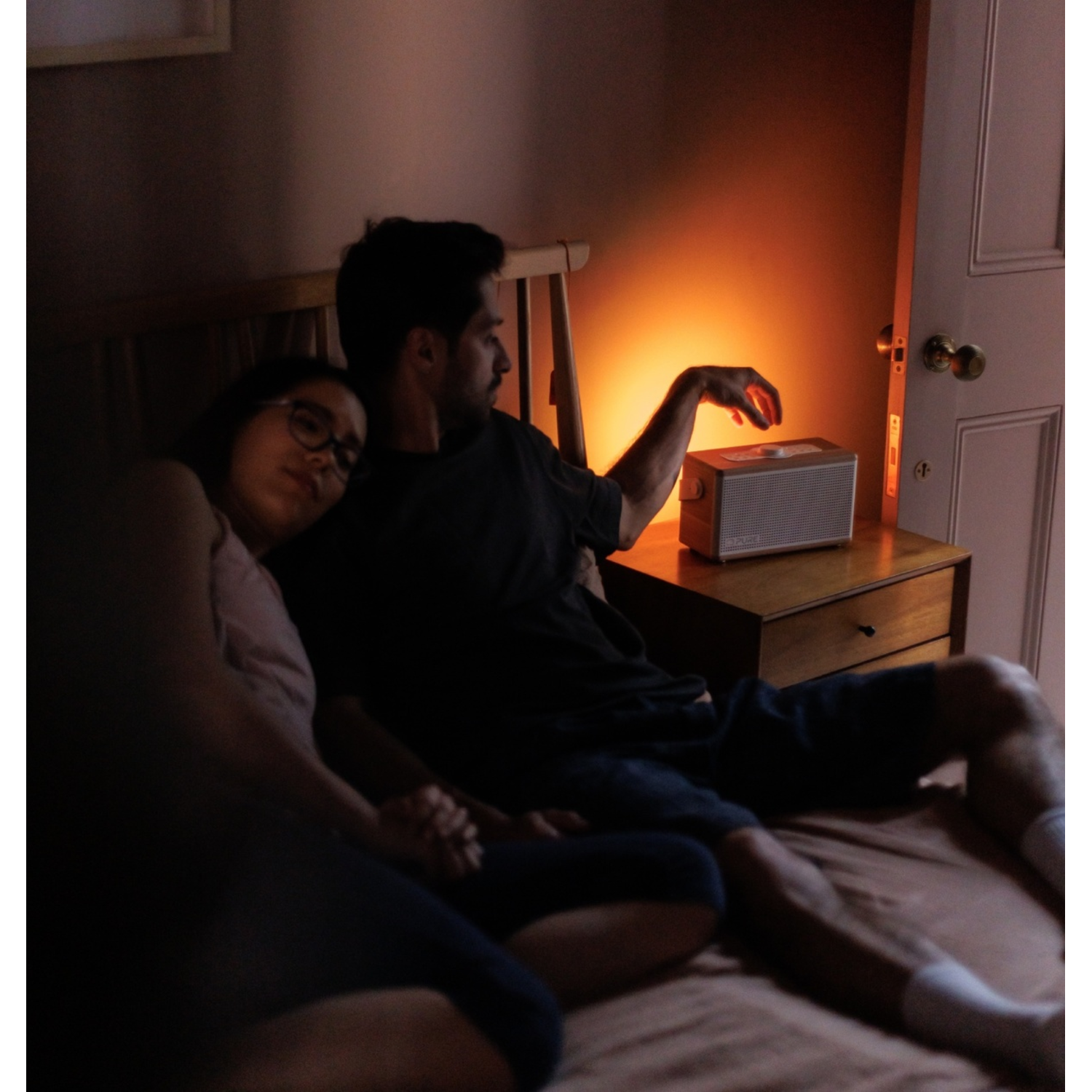 Paar im Bett, hält Hände. Mann greift nach Lautsprecher. Weiches Licht erhellt den Raum. Rechts ist eine Tür.