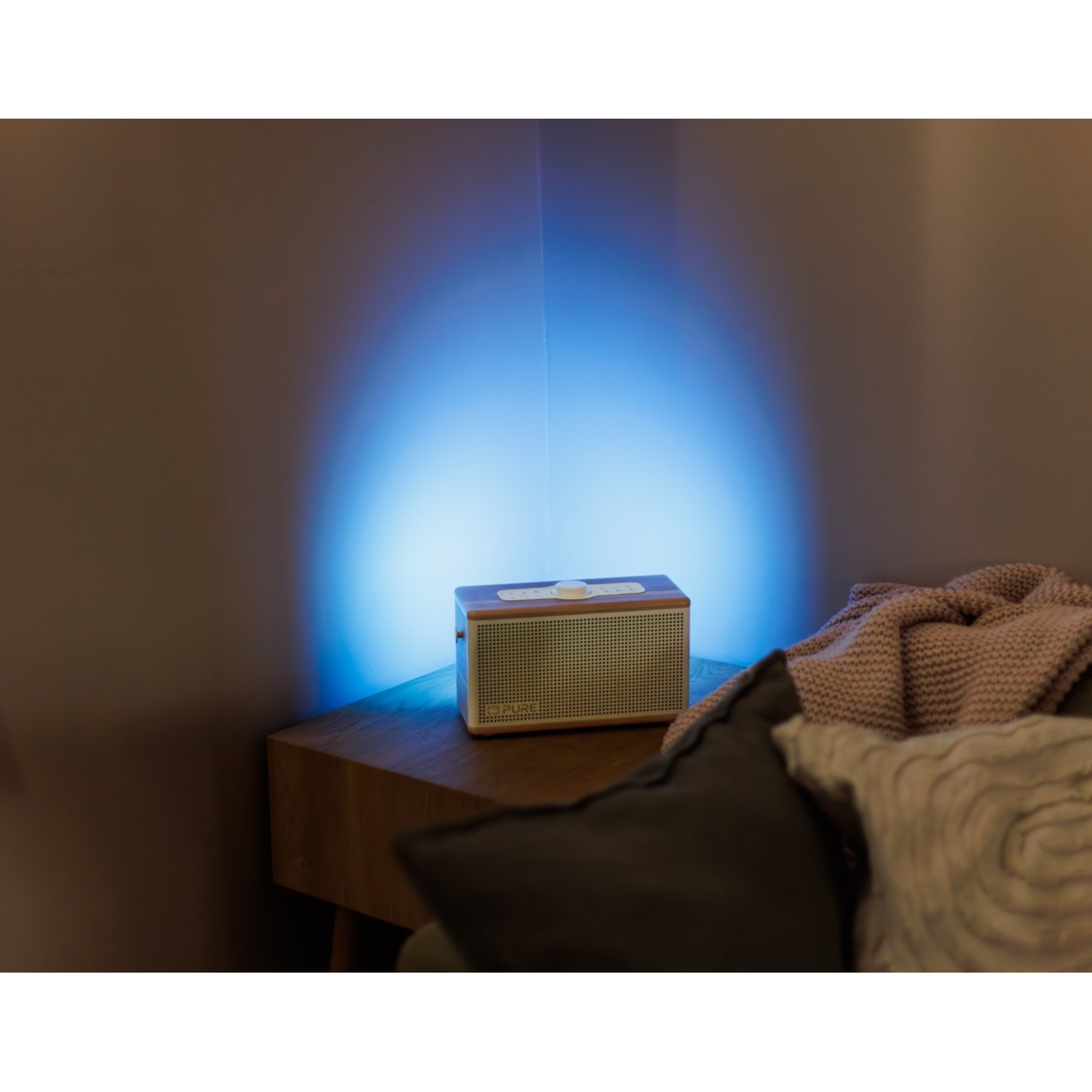 Ein Lautsprecher auf einem Nachttisch. Blaues Licht scheint auf die dahinterliegende Wand. Kissen und eine Decke sind im Vordergrund.