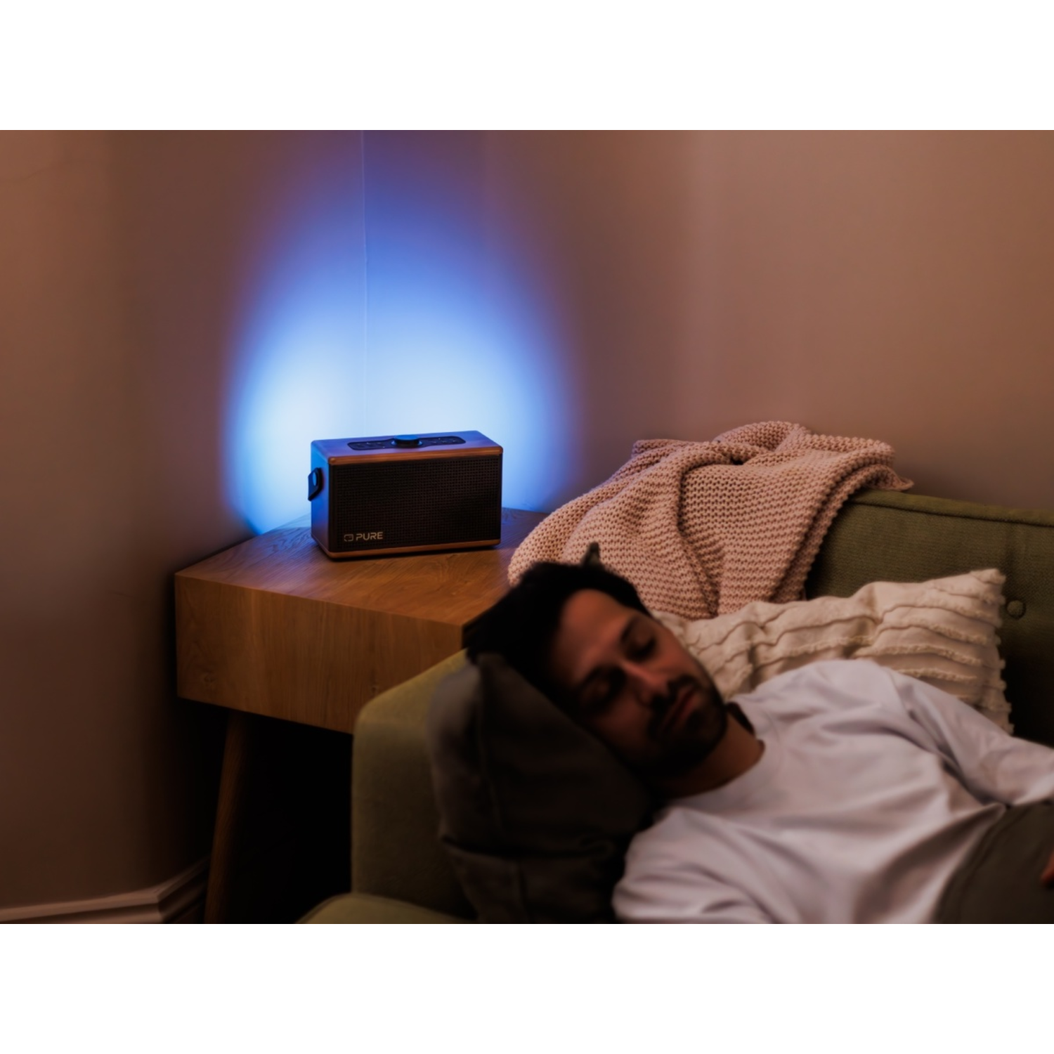 Ein Mann schläft auf einem grünen Sofa mit Kissen und Decke. Auf einem Holztisch steht ein Lautsprecher mit blauem Licht.