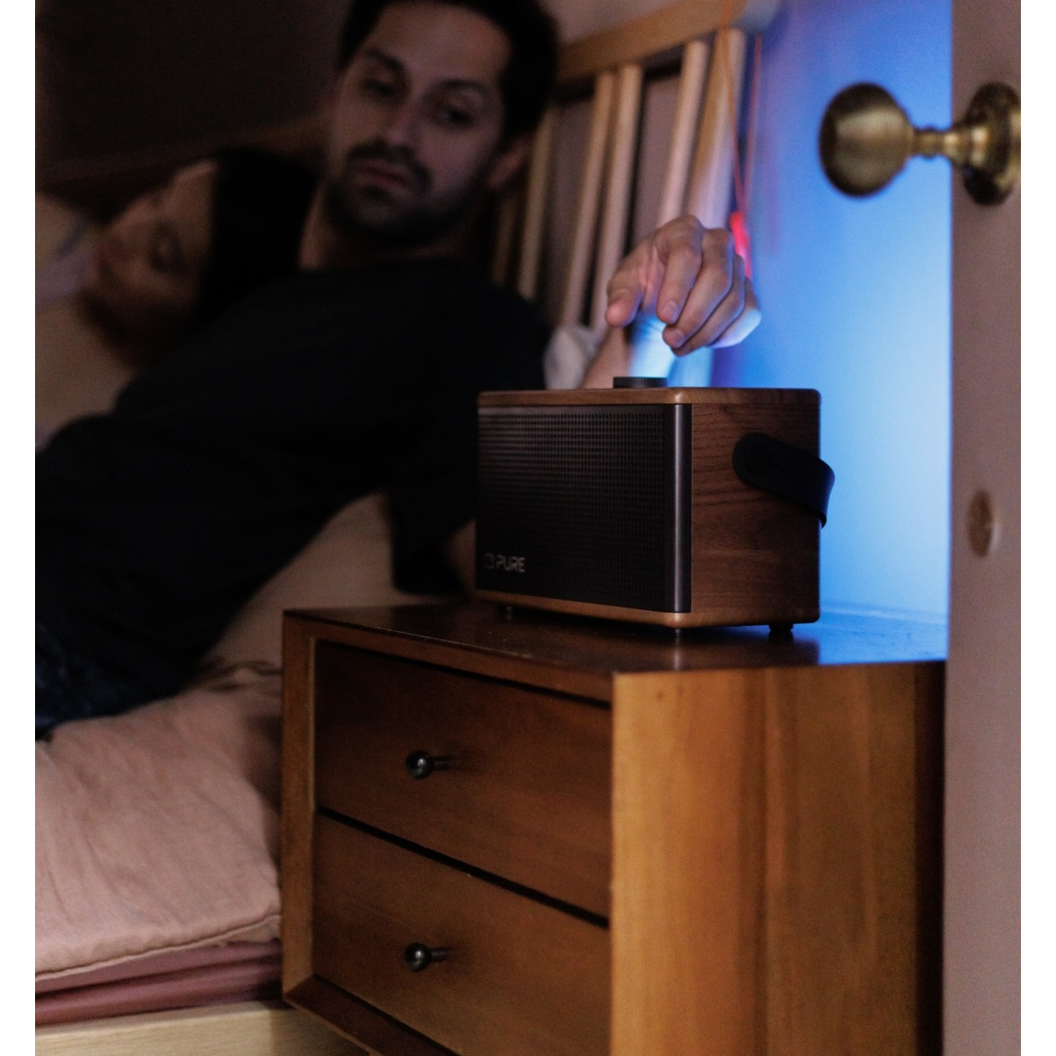 Ein Mann greift nach einem Lautsprecher auf einem Nachttisch neben einem Bett mit einer schlafenden Person. Im Hintergrund ist eine Tür geöffnet.