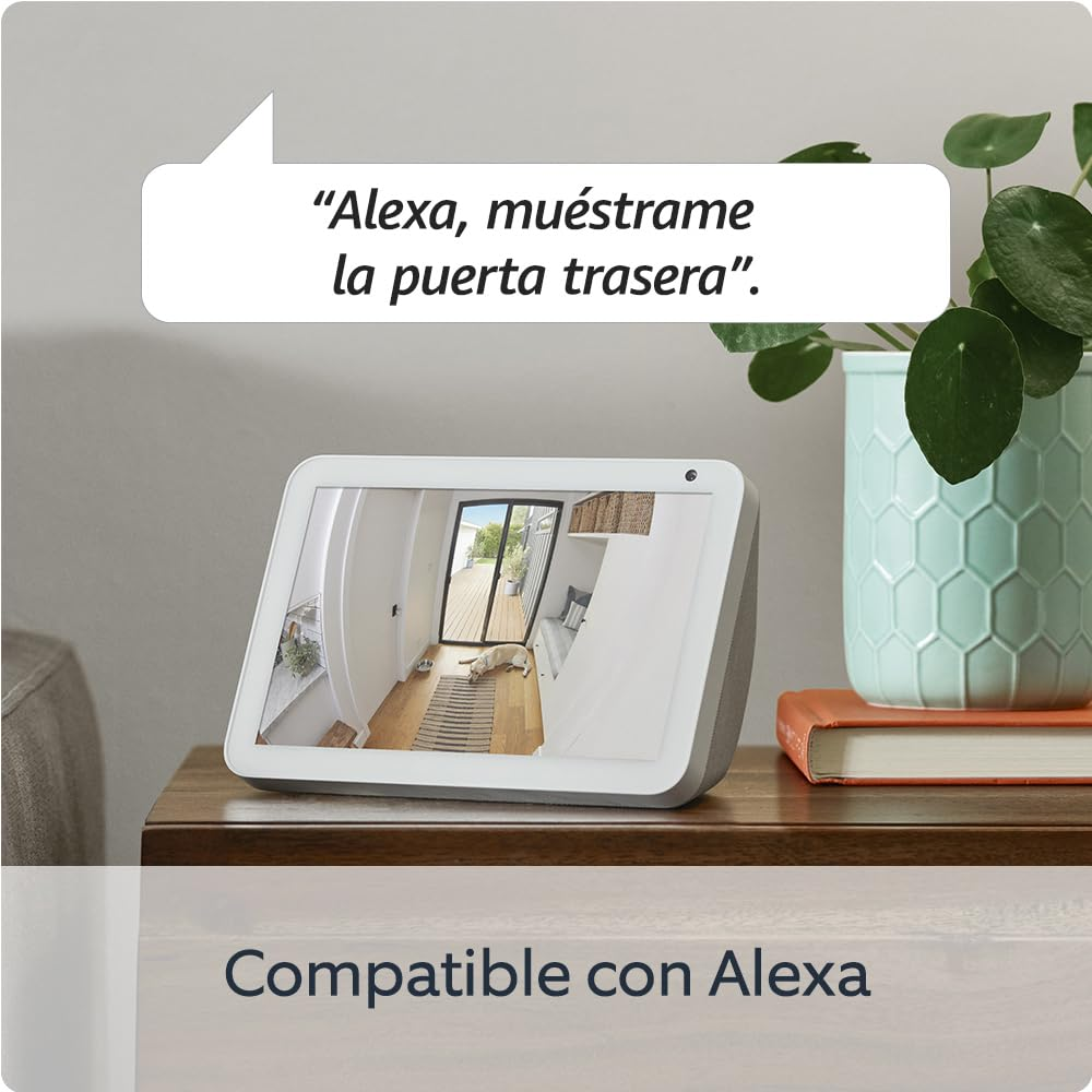 Un dispositivo Alexa muestra una vista de la puerta. El texto dice: 'Alexa, muéstrame la puerta trasera'. Hay una planta, un libro y el texto 'Compatible con Alexa'.