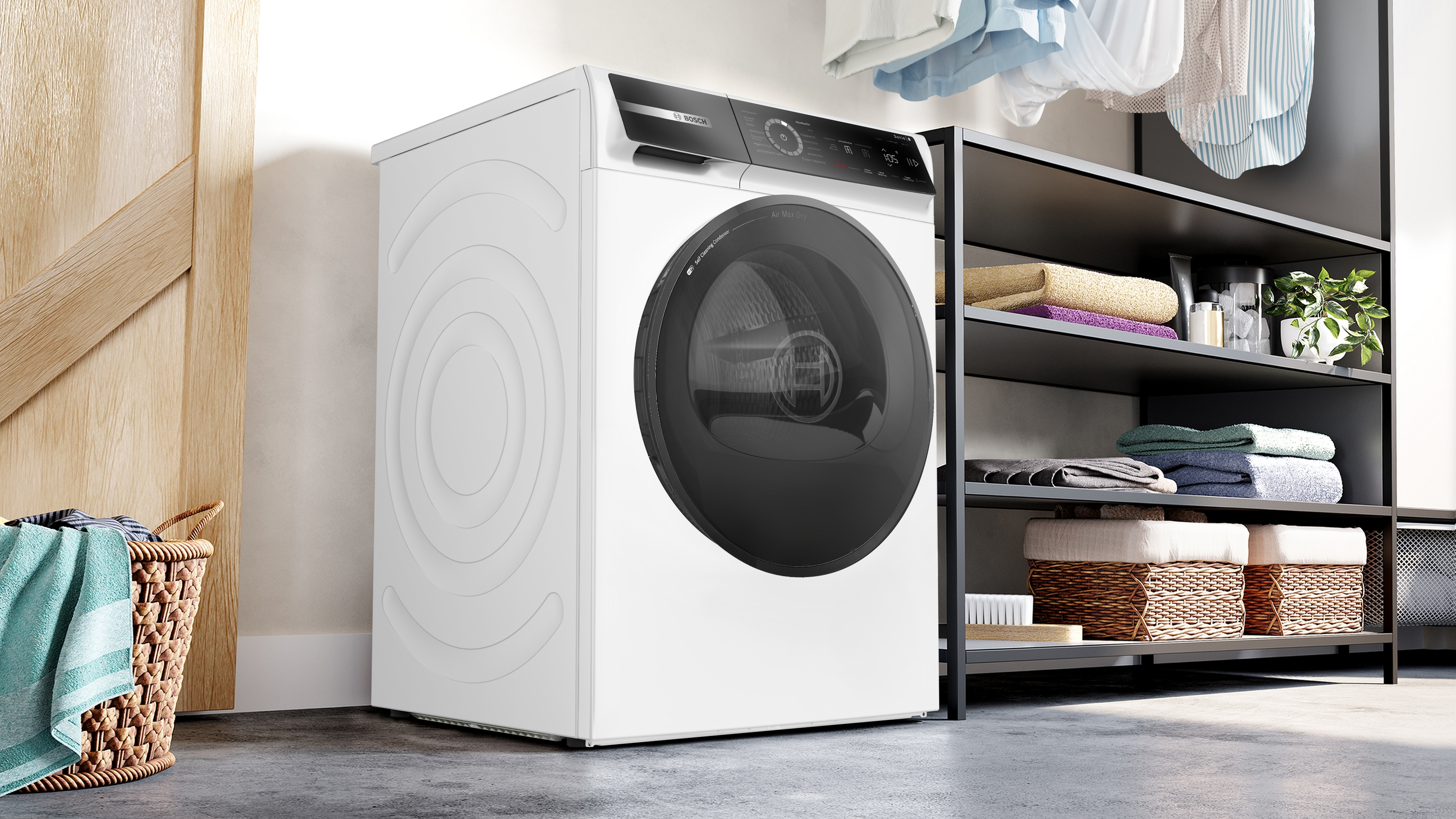 Ein weißer Bosch-Trockner steht neben einem Regal mit Handtüchern. Im Vordergrund steht ein Korb mit Handtüchern. Kleidung hängt darüber.