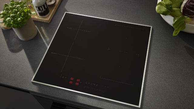 Czarna płyta indukcyjna ze srebrną ramą na szarym blacie. Zioła i olej obok.