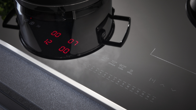 Czarny garnek na czarnej płycie kuchennej. Czerwony wyświetlacz cyfrowy pokazuje 12:00, 00:07 i 00:00. Płyta ma ustawienia temperatury i elementy sterujące.
