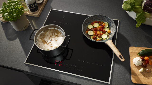 Scena kuchenna z czarną płytą indukcyjną. Garnek gotuje się z jednej strony, patelnia z warzywami z drugiej.