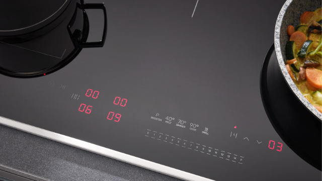 Czarna płyta grzewcza z czerwonymi wyświetlaczami cyfrowymi. Patelnia z warzywami po prawej. Garnek po lewej.