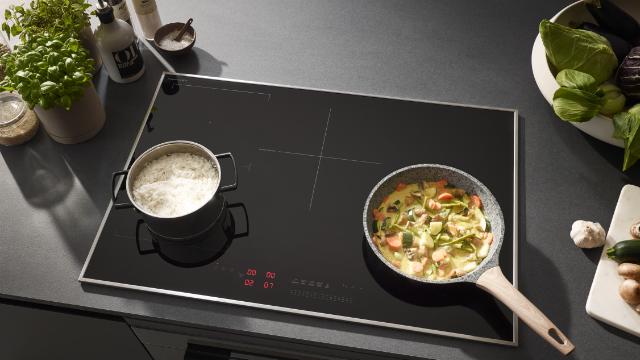 Czarna płyta indukcyjna z ryżem w garnku i warzywami na patelni. Inne składniki po bokach.