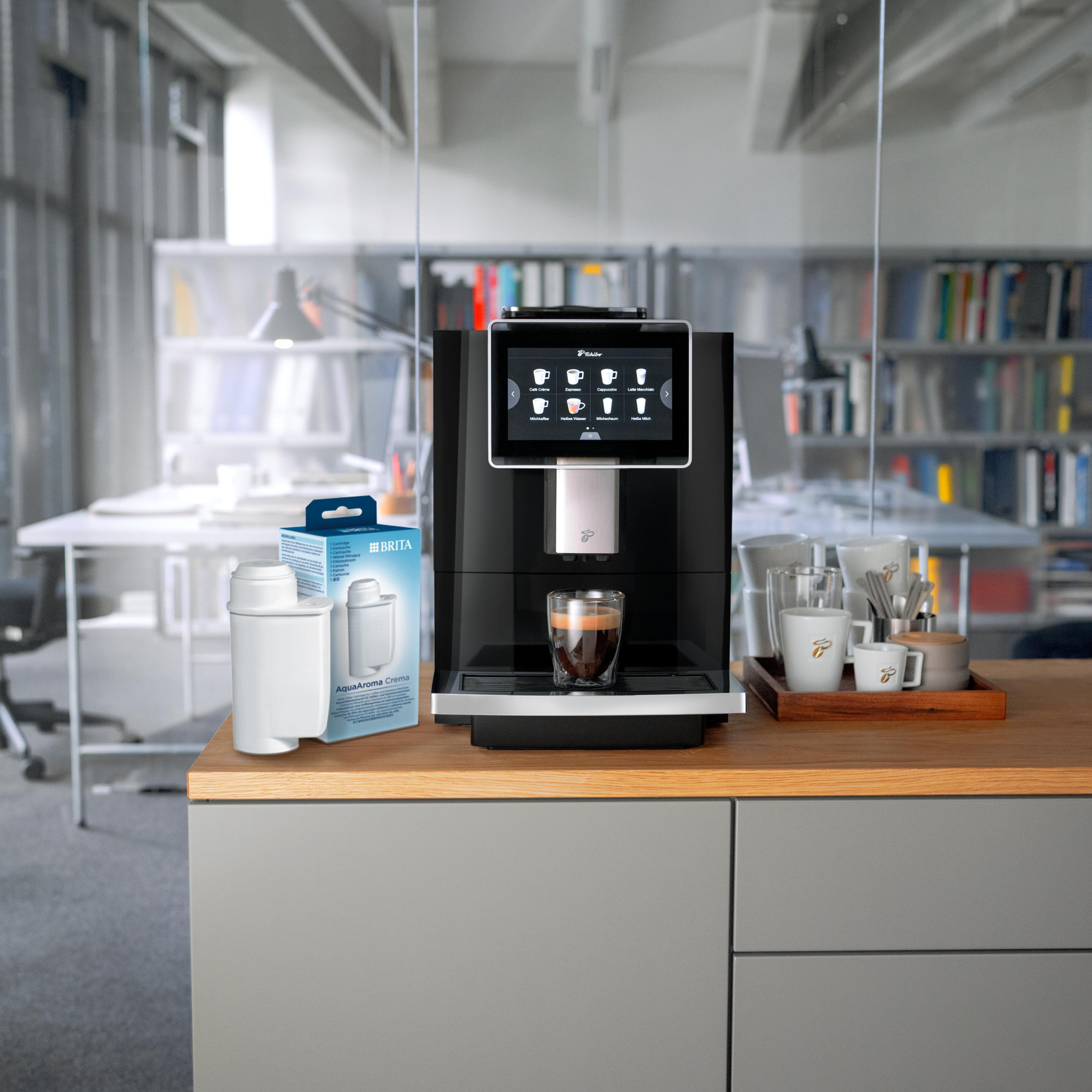 Kaffeemaschine mit Espresso, Kaffeetassen und Wasserfilter auf einer Holztheke in einem Büro.