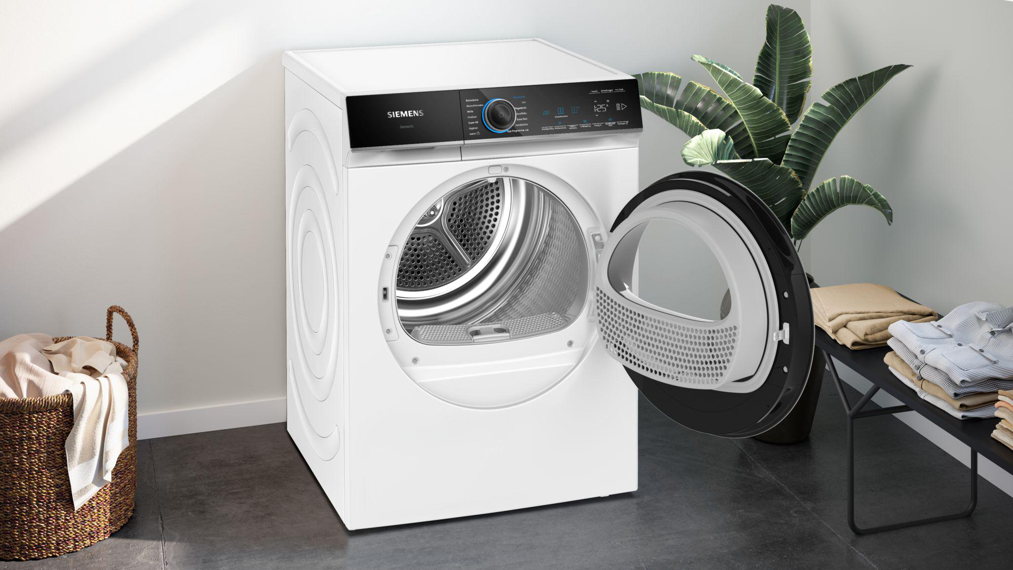 Weißer Siemens-Trockner mit offener Tür, Kleidung im Inneren. Wäschekorb und Pflanzen an der Seite. Schwarzes Regal mit gefalteter Kleidung.