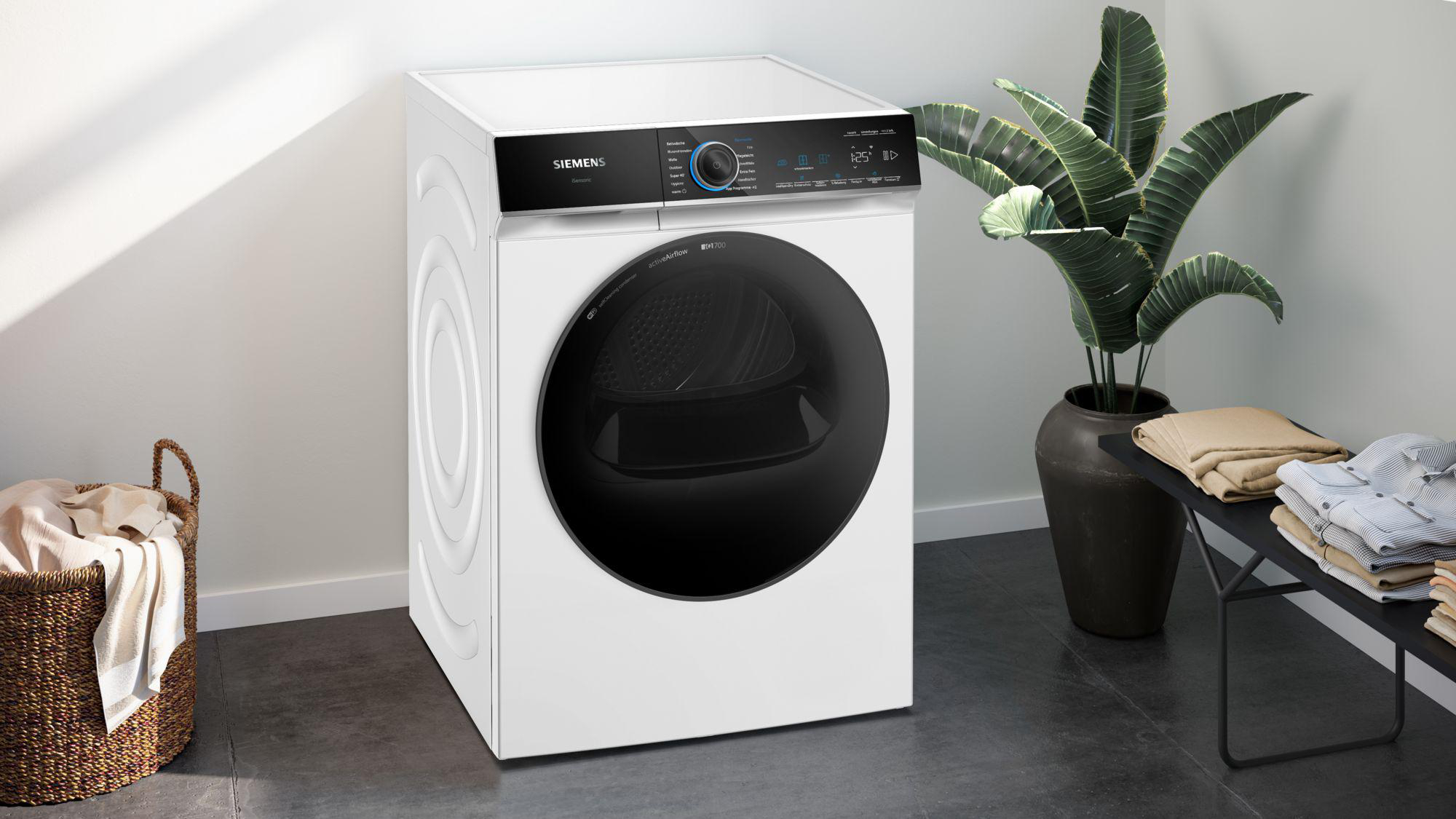 Ein weißer Siemens-Trockner. Links ein Korb mit Kleidung. Rechts eine Pflanze und eine Bank mit gefalteter Kleidung.