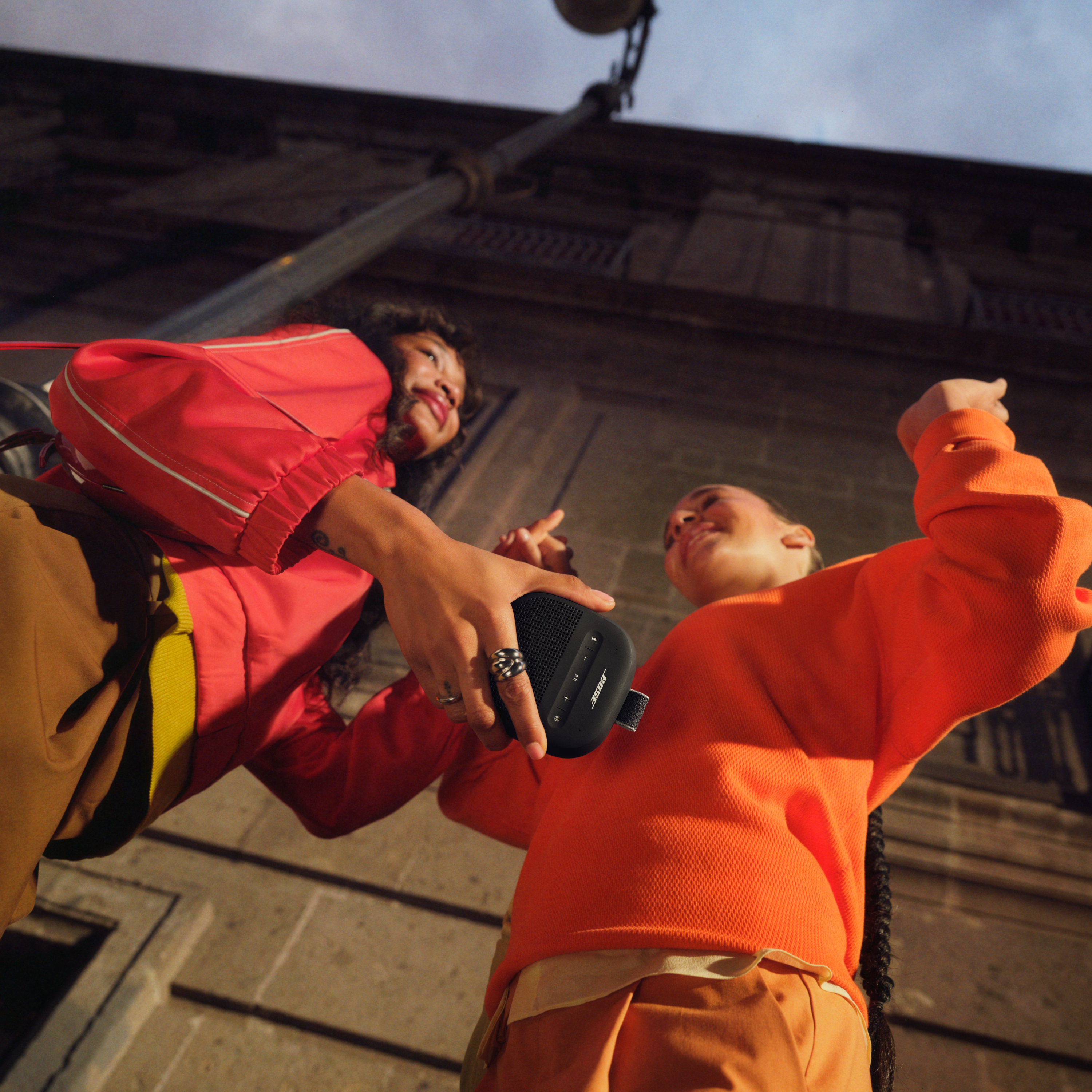 Zwei Frauen halten einen schwarzen Lautsprecher. Eine trägt eine rote Jacke, die andere einen orangefarbenen Pullover. Im Hintergrund ein Gebäude und eine Straßenlaterne.