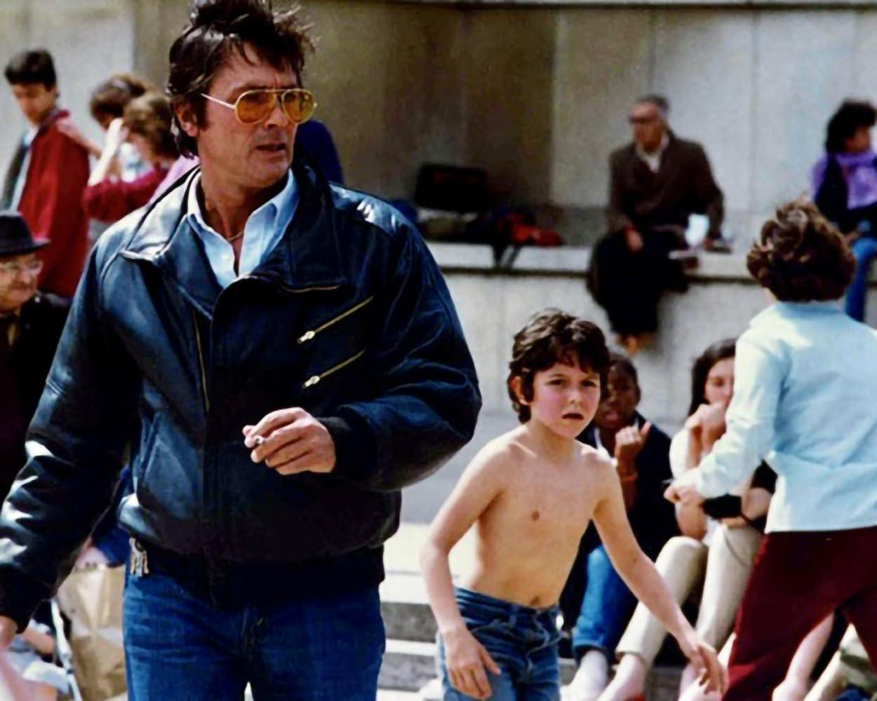 Mann mit Sonnenbrille und Lederjacke mit einem oberkörperfreien Jungen und anderen Leuten im Hintergrund.