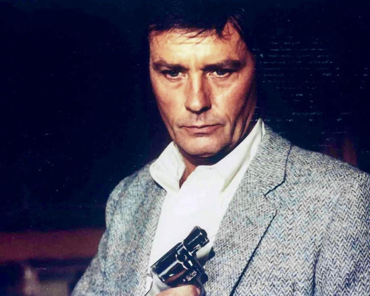 Ein Mann in einem grauen Blazer und weißem Hemd hält eine Waffe. Er hat dunkle Haare und einen ernsten Gesichtsausdruck. Dunkler Hintergrund.