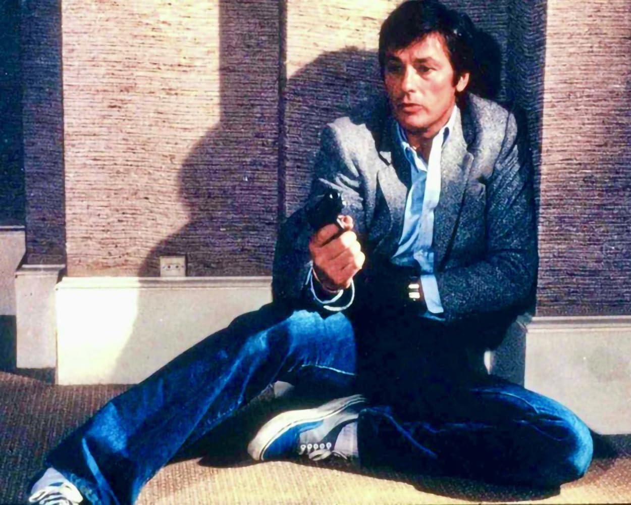 Ein Mann in Blazer und Jeans hält eine Pistole, sitzt an einer strukturierten Wand. Er trägt Turnschuhe. Schatten deuten auf einen sonnigen Tag hin.